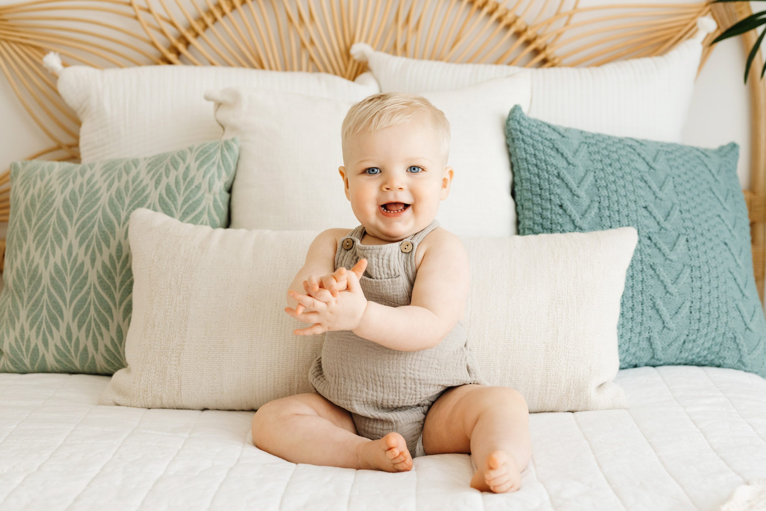 a little boy sitting on a bed and laughing as he claps his hands together during a first birthday photography session