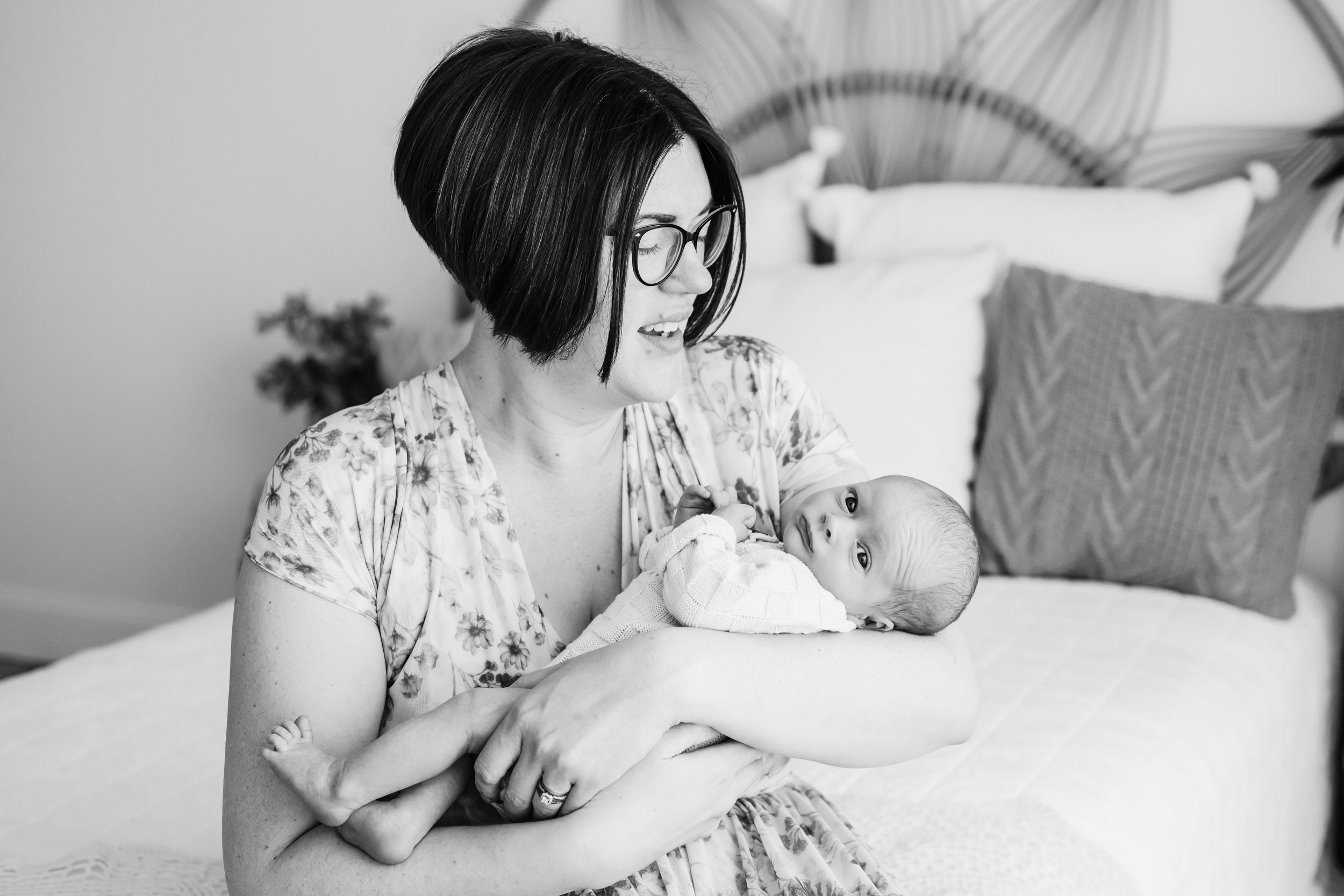 a black and white picture of a mom sitting on a bed with her baby boy cradled in her arms as she smiles down at him during a newborn photos session