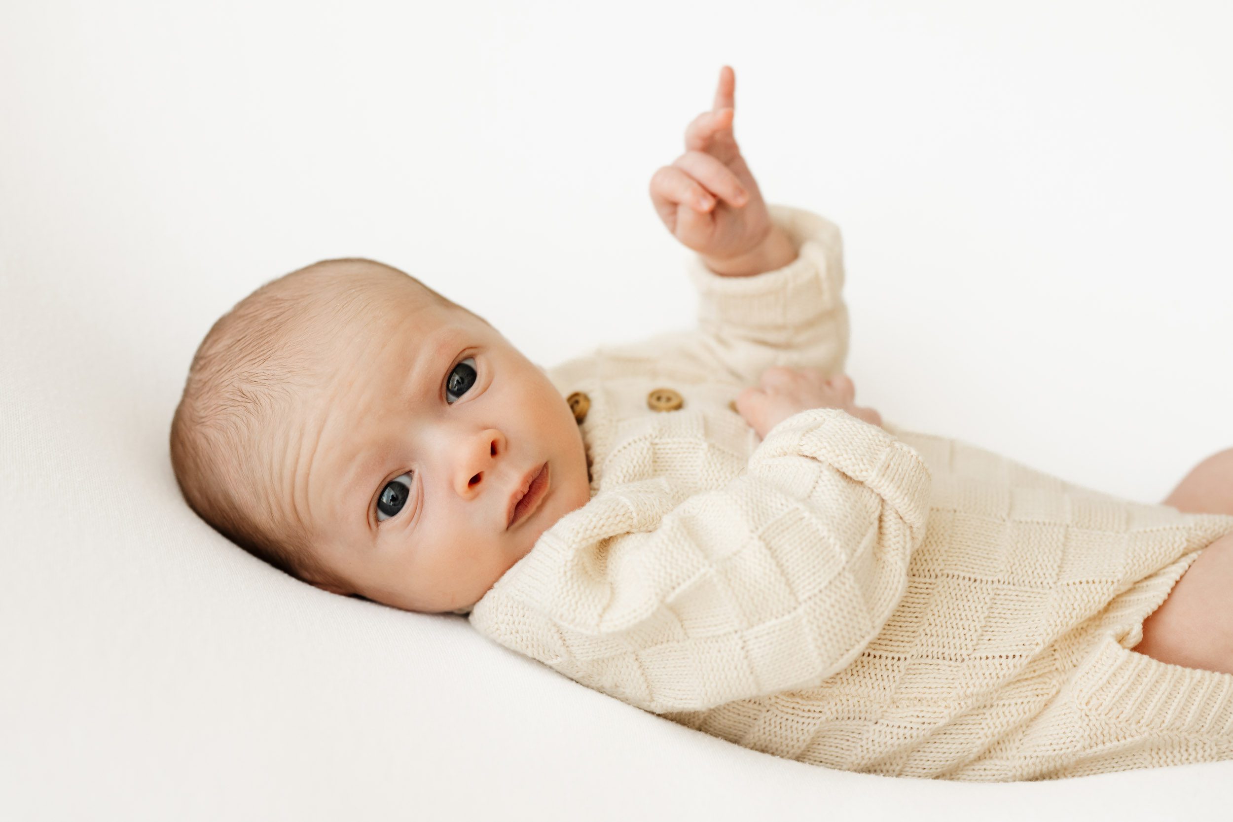 a baby boy wearing a cream colored knit onesie laying on his back and gazing right at the camera as he lifts his hand up into the air during a newborn photos session