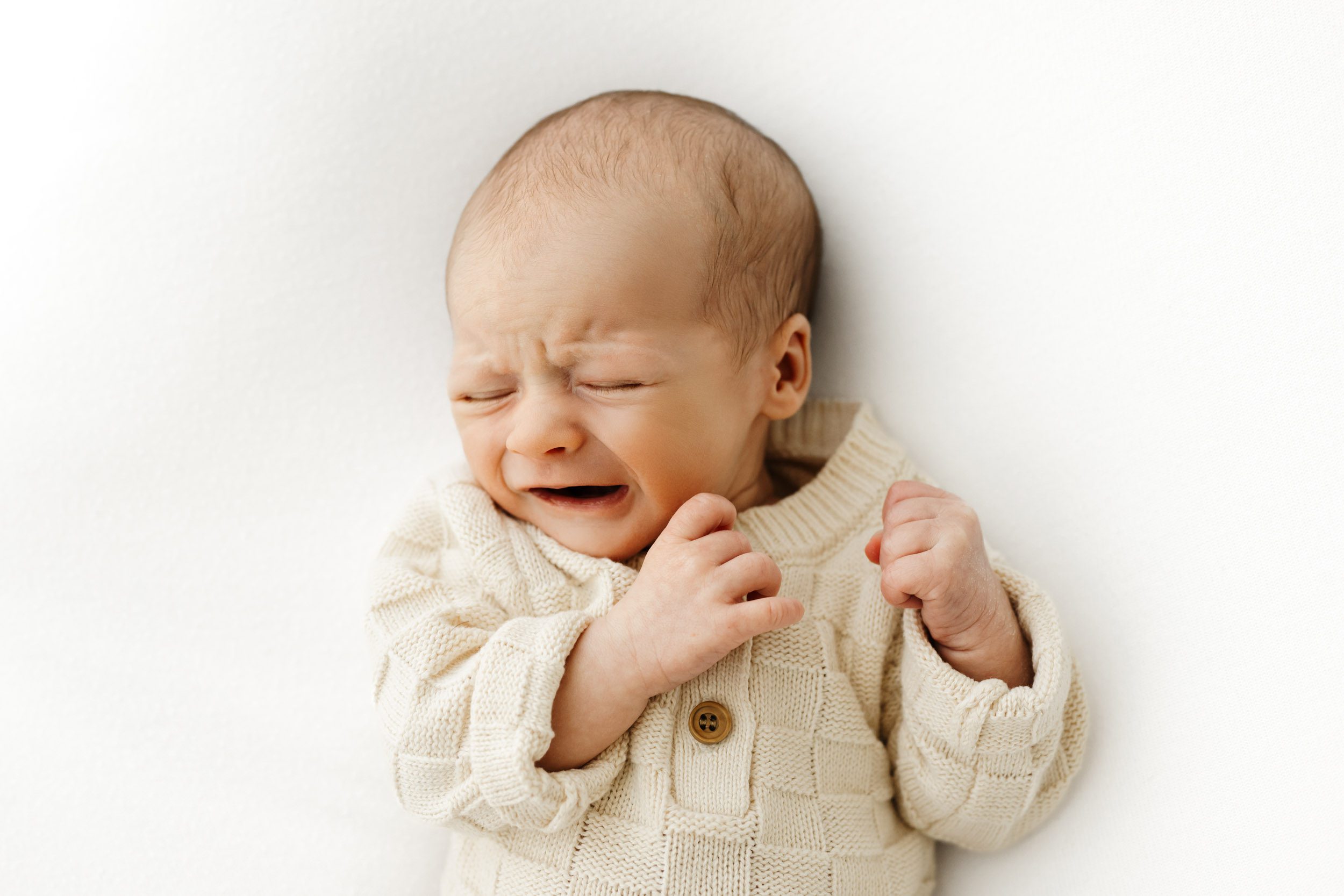 a baby boy wearing a cream knit onesie laying on a white backdrop and crying during a newborn photos session