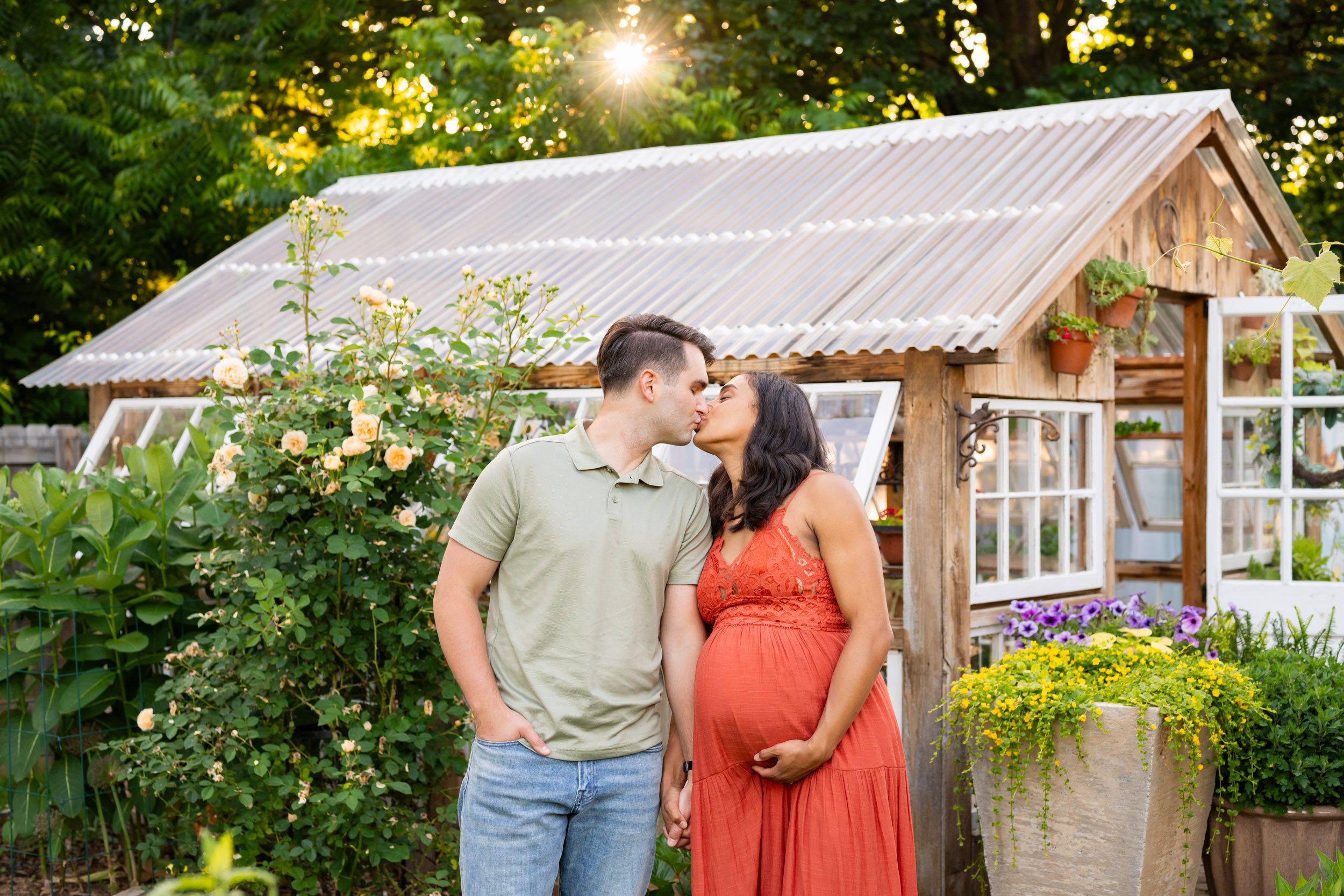 expecting parents standing in a garden in front of a greenhouse kissing each other while the sunset shines through the trees in the background during a West Chester maternity photoshoot