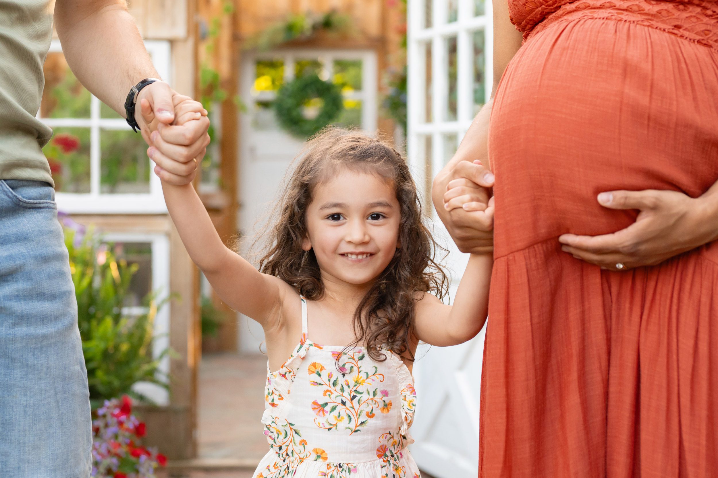 a young girl wearing a floral dress holding her parents' hands and smiling directly at the camera during a West Chester maternity photoshoot