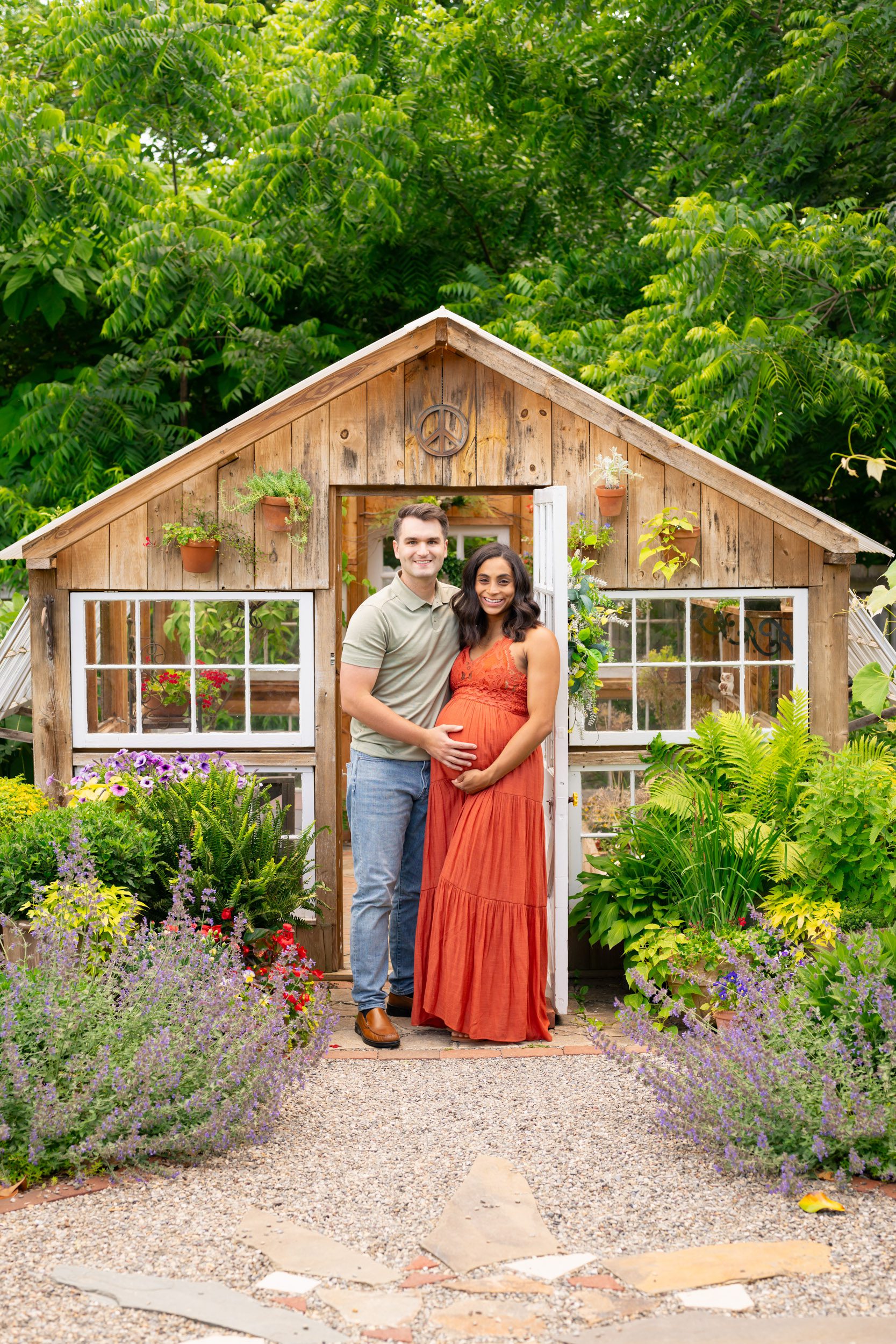 expecting parents standing in a garden in front of a greenhouse as they touch mom's belly and smile at the camera during a West Chester maternity photoshoot
