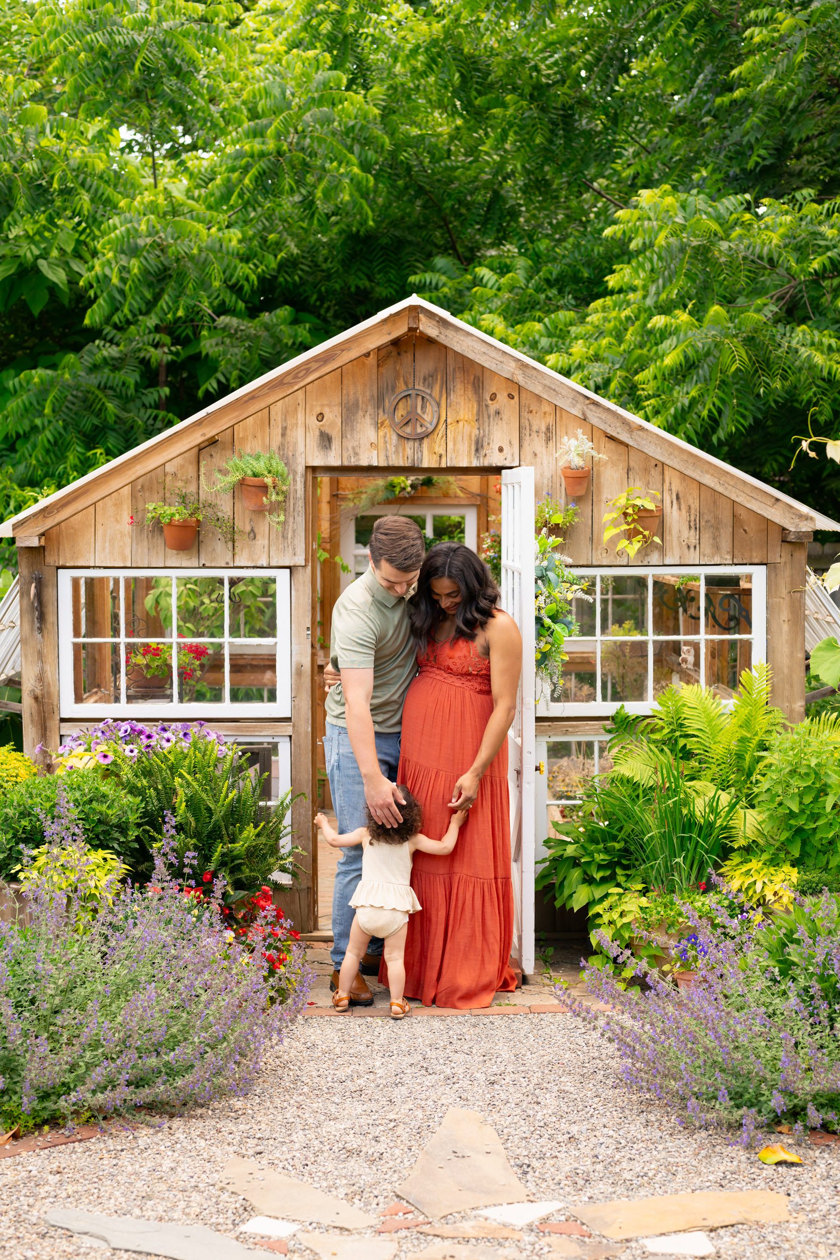 expecting parents standing in a garden in front of a greenhouse as their young daughter runs up to hug them and they reach down to touch her head during a West Chester maternity photoshoot