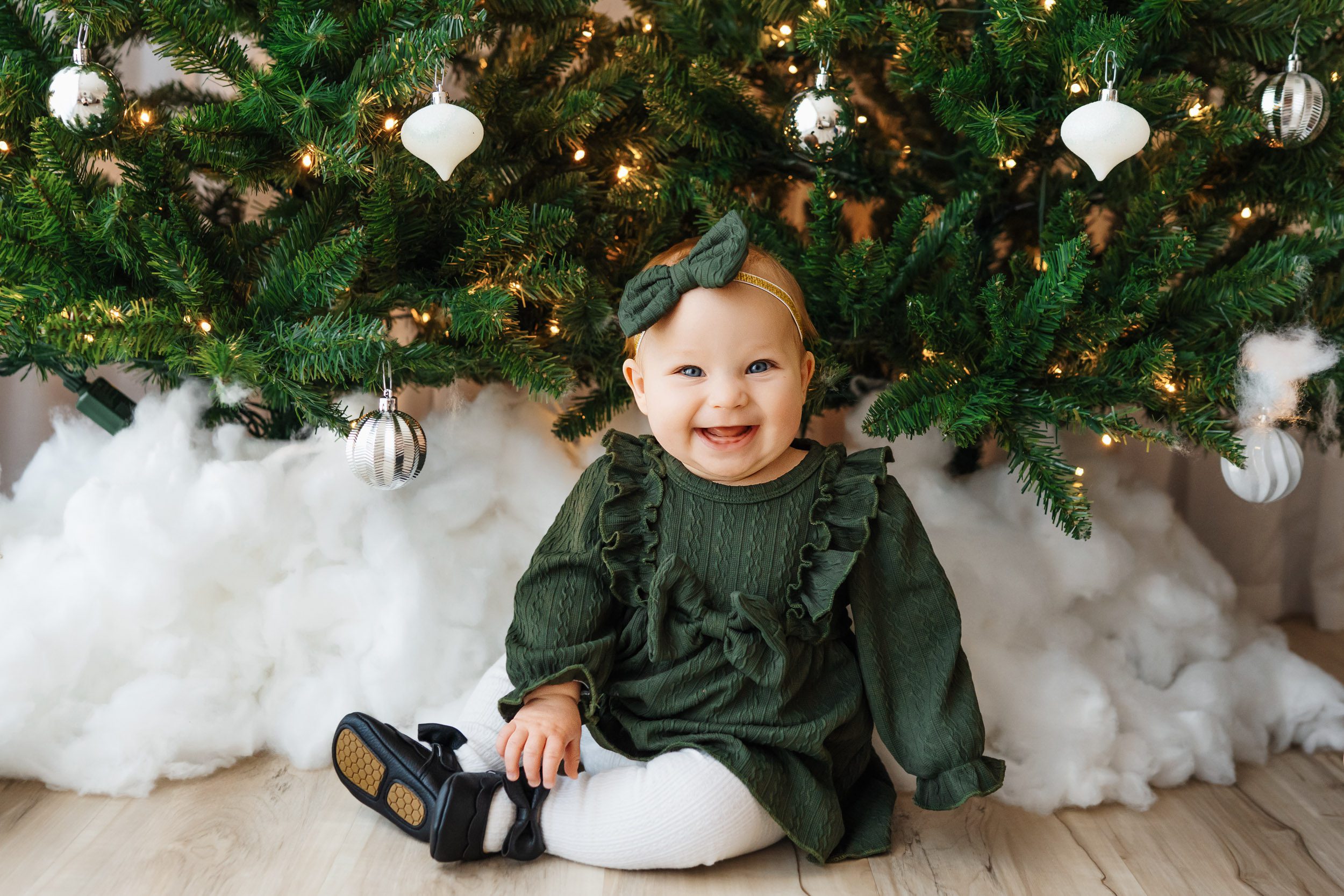 a little girl sitting on the floor in front of a Christmas tree decorated with white and silver ornaments as she looks at the camera with a huge smile on her face during a Christmas mini session
