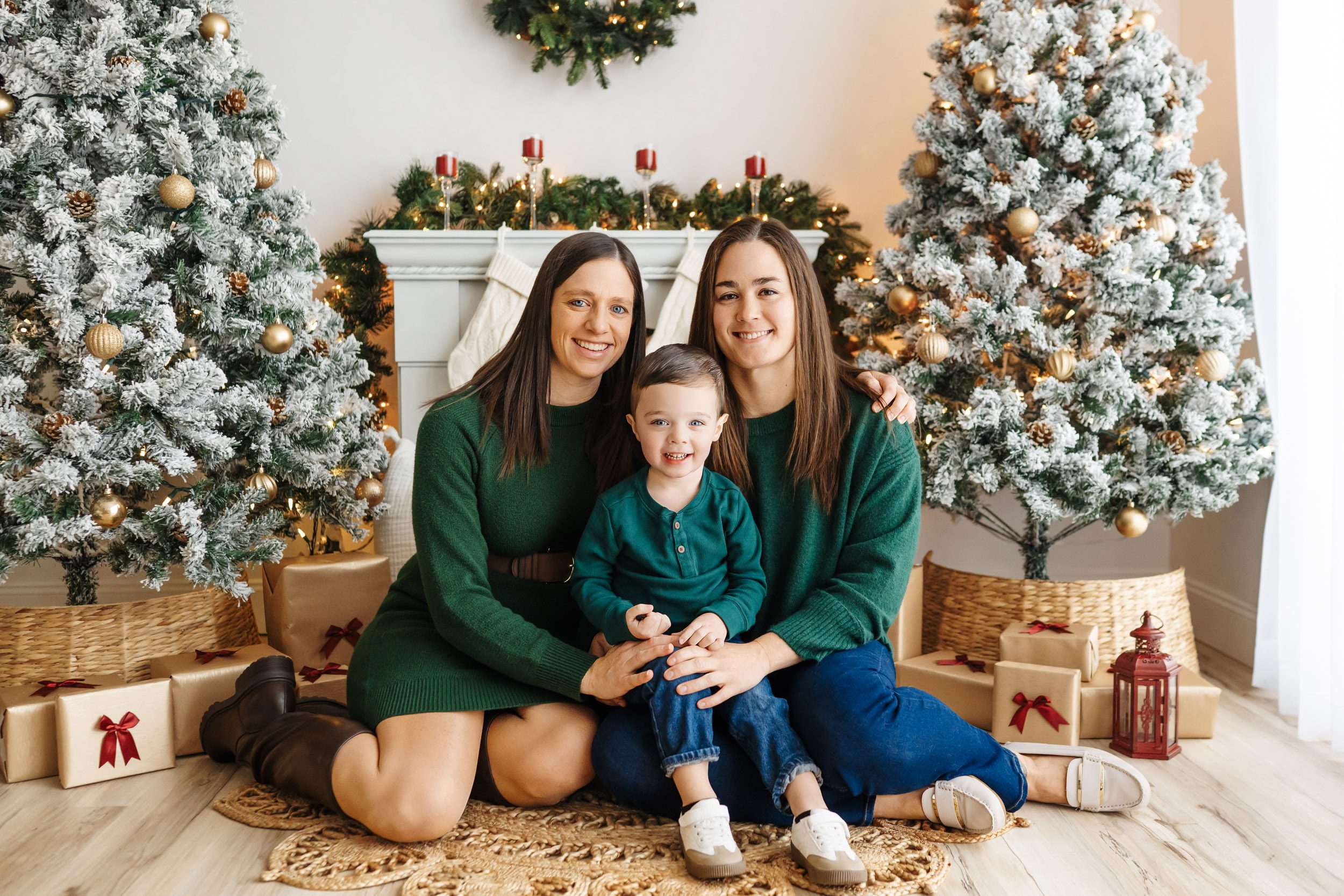 two moms sitting on the floor with their son on their lap surrounded by snow-flocked Christmas trees and holiday decor as they all smile at the camera during a Christmas mini session