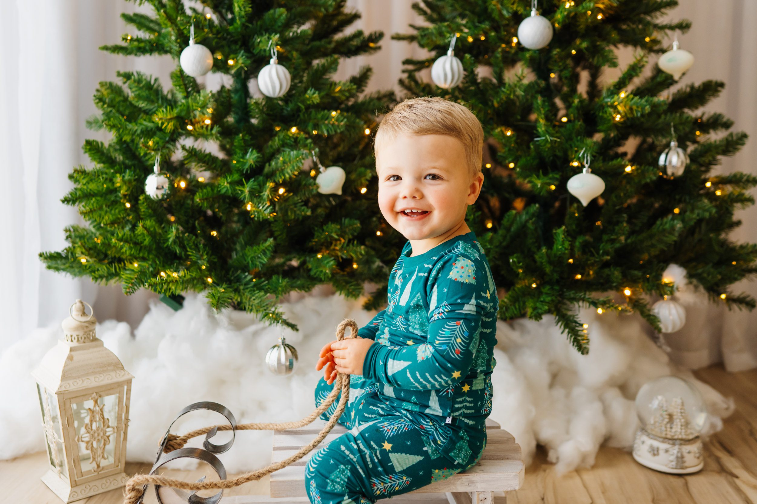 a brother and sister sitting on a white loveseat surrounded by snow-flocked Christmas trees and holiday decor as they both smile at the camera during a Christmas mini session