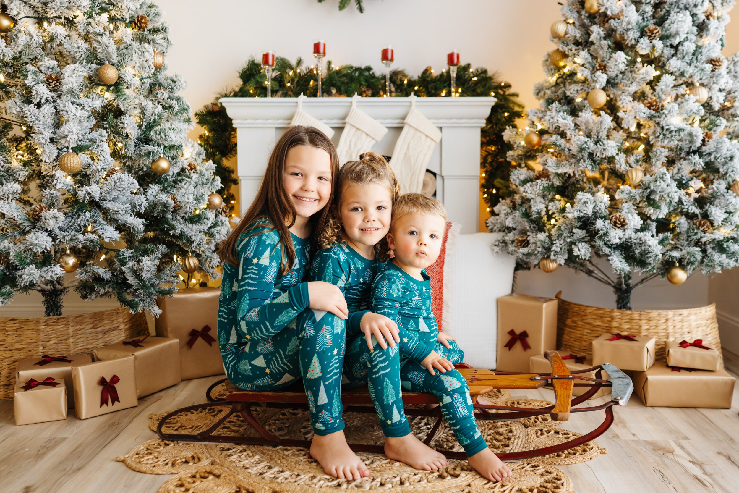 three young kids wearing matching green holiday pajamas sitting on a red antique sled surrounded by snow-flocked Christmas trees and holiday decor as they all smile at the camera during a Christmas mini session