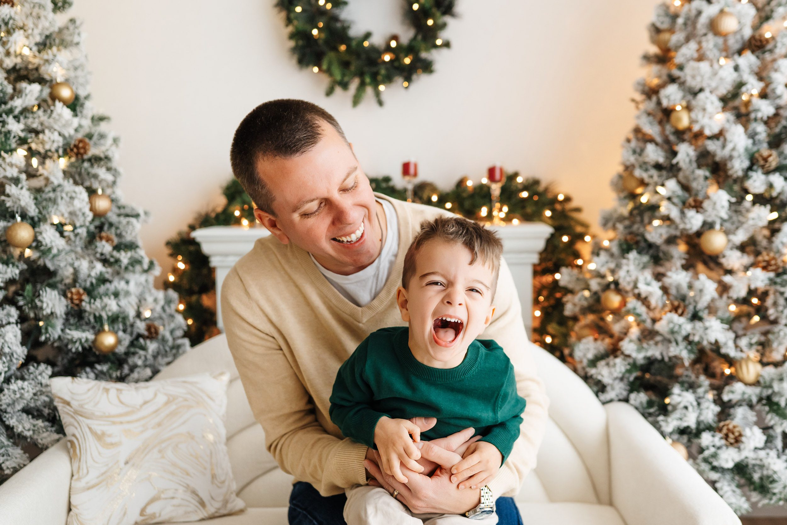 a young boy sitting on his dad's lap with a huge smile on his face surrounded by snow-flocked Christmas trees and holiday decor during a Christmas mini session