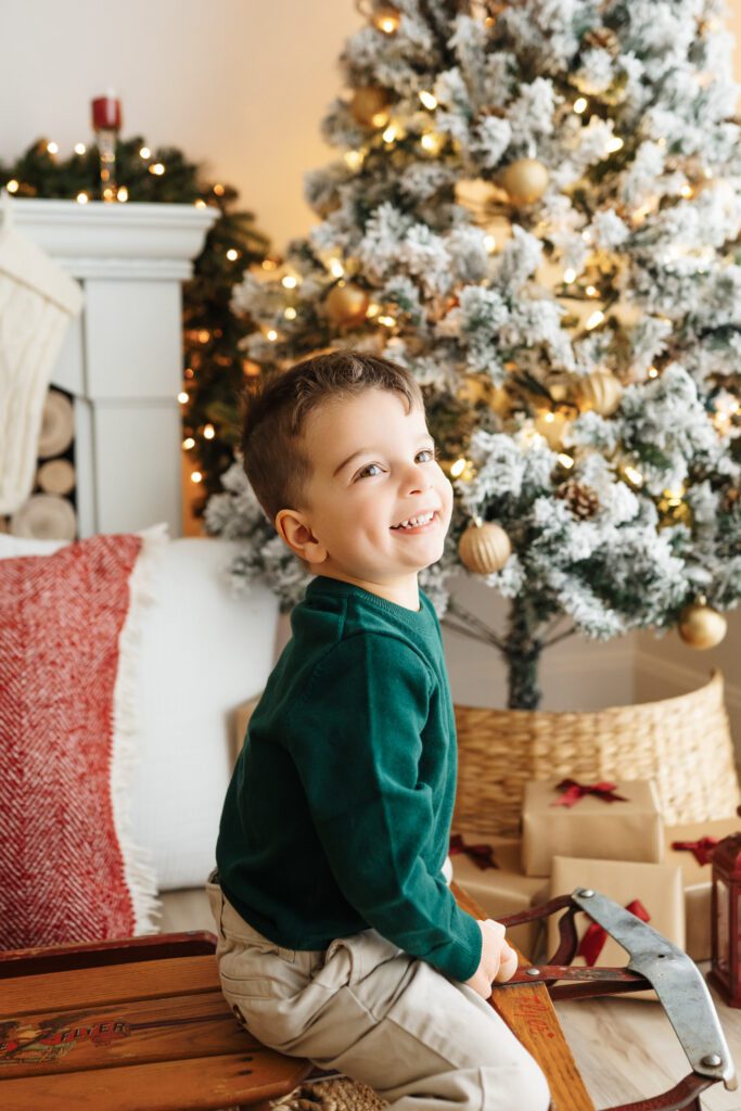 a young boy sitting on an antique sled surrounded by snow-flocked Christmas trees and holiday decor as he looks up with a huge smile on his face during a Christmas mini session