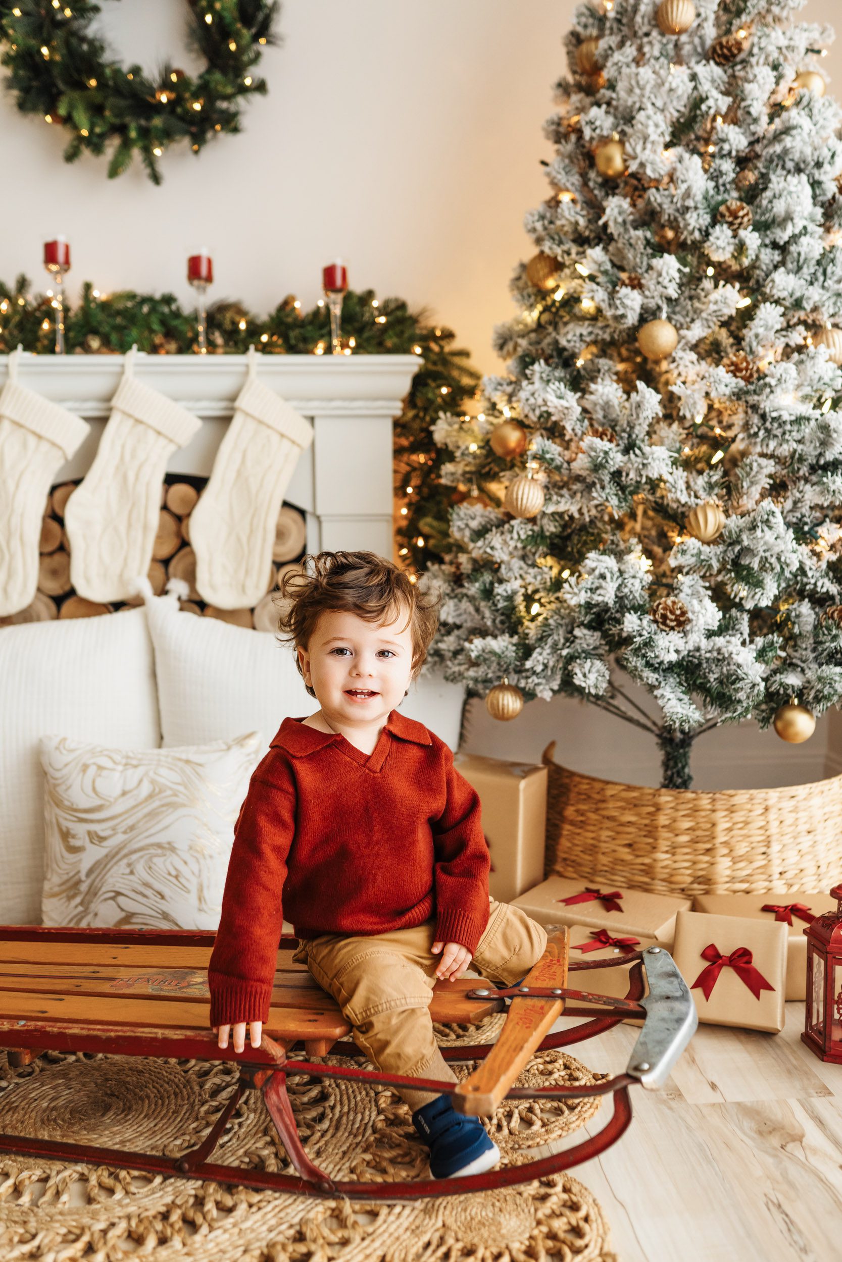 a young boy wearing a red sweater sitting on an antique sled surrounded by snow-flocked Christmas trees and holiday decor as he smiles at the camera during a Christmas mini session