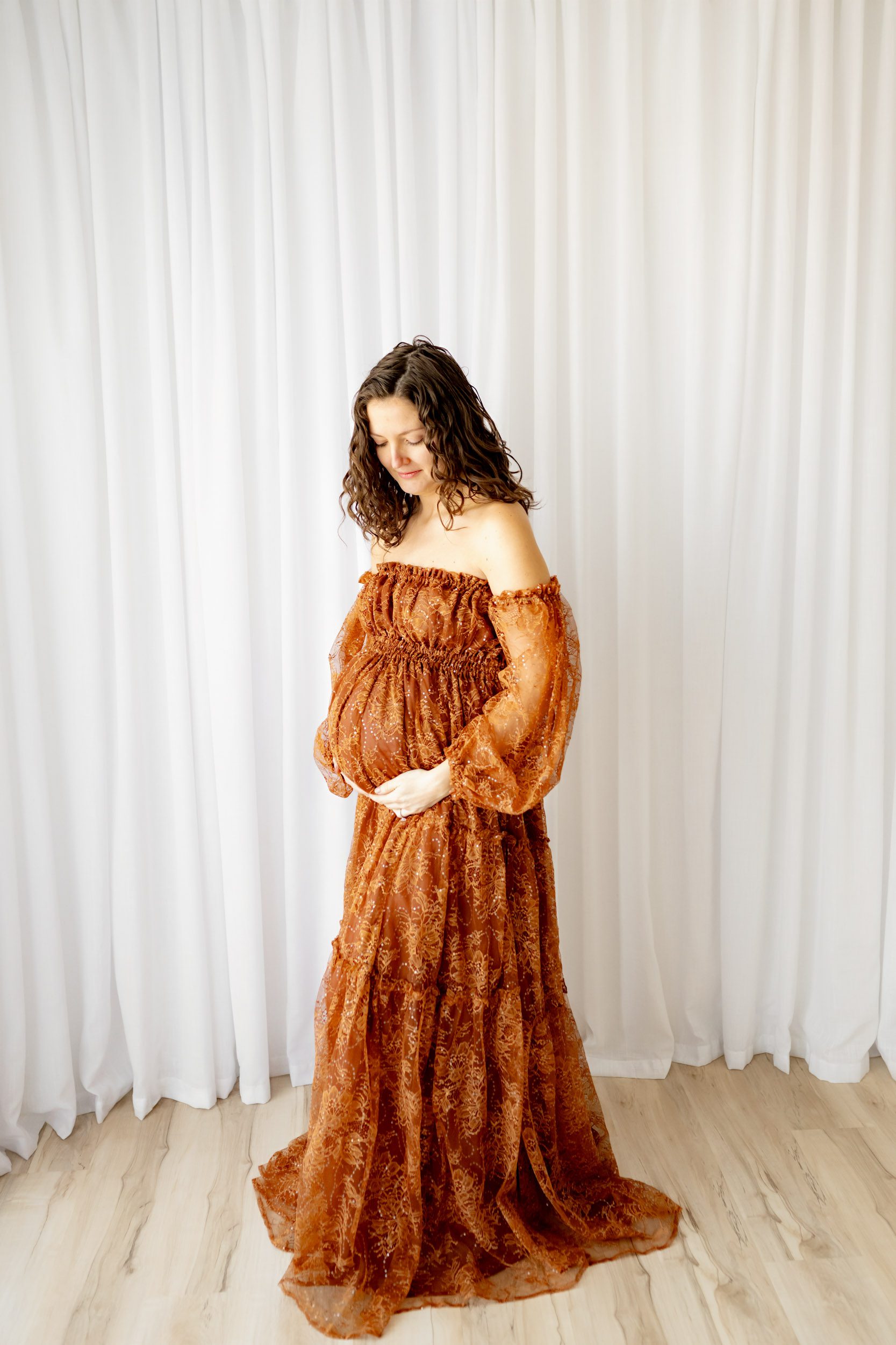 an expecting mother wearing a rust colored lace maternity dress standing in front of a white curtain and cradling her belly