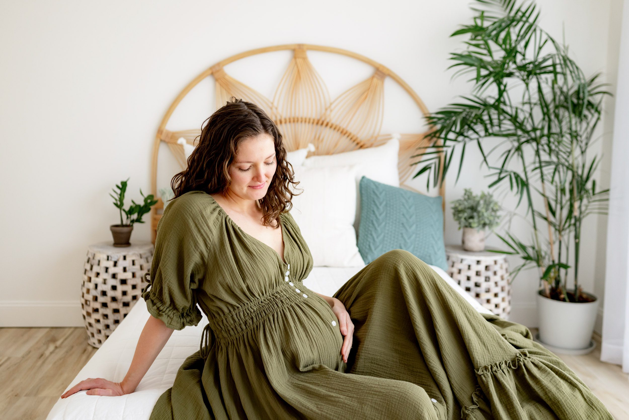 an expecting mother wearing an olive green maternity dress sitting in on a bed and smiling down at her belly