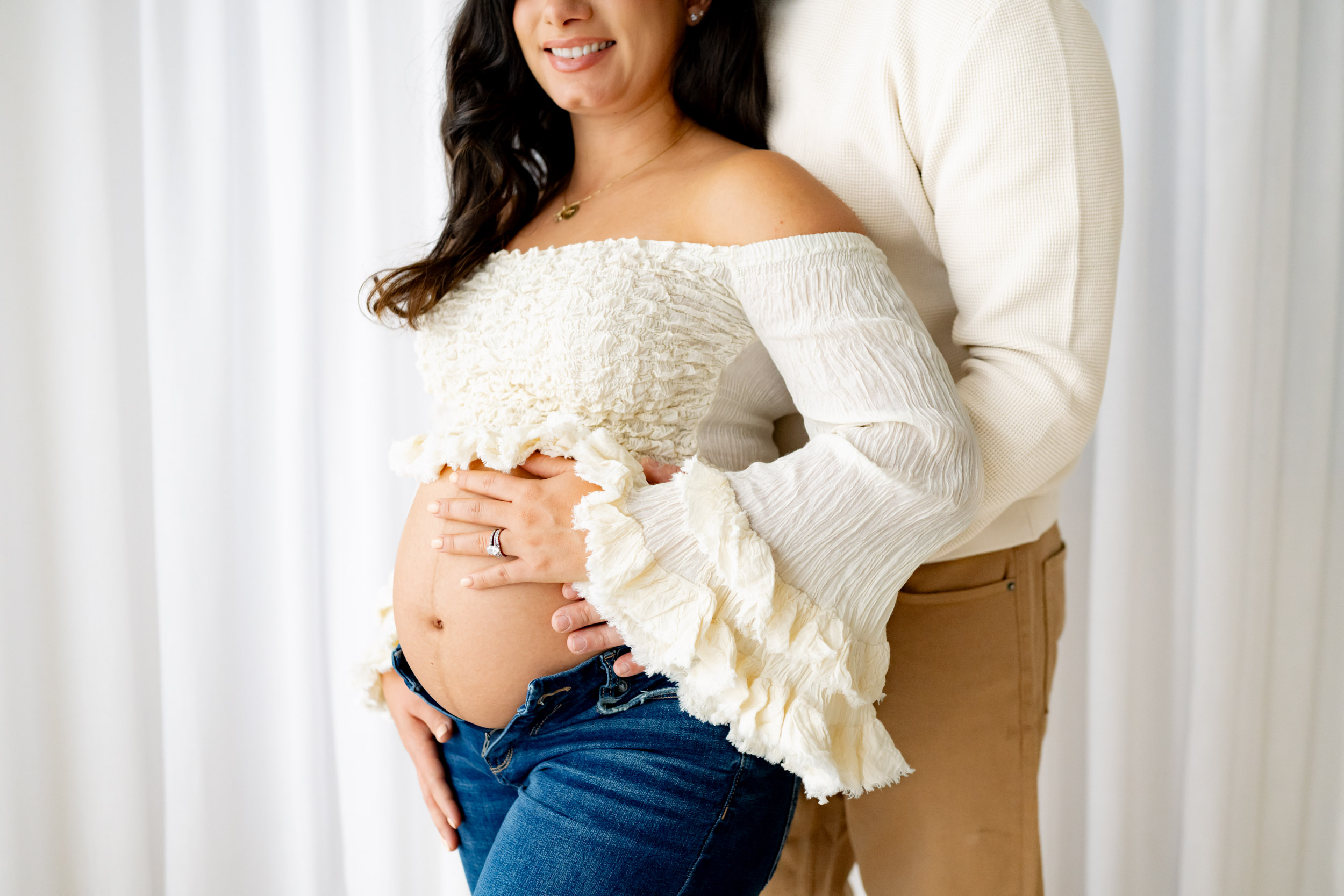 a close-up image of an expecting mother donning a textured top with flowing sleeves and jeans as her partner embraces her from the back while both place their hands on her exposed belly during a lifestyle maternity photoshoot