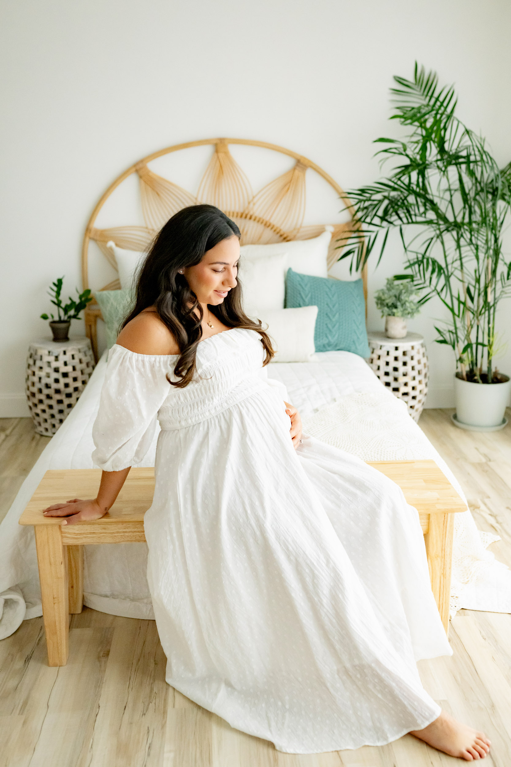 an expecting mother dressed in a flowing white off-the-shoulder gown seated on a bench before a bed, smiling affectionately at her belly during a lifestyle maternity photoshoot