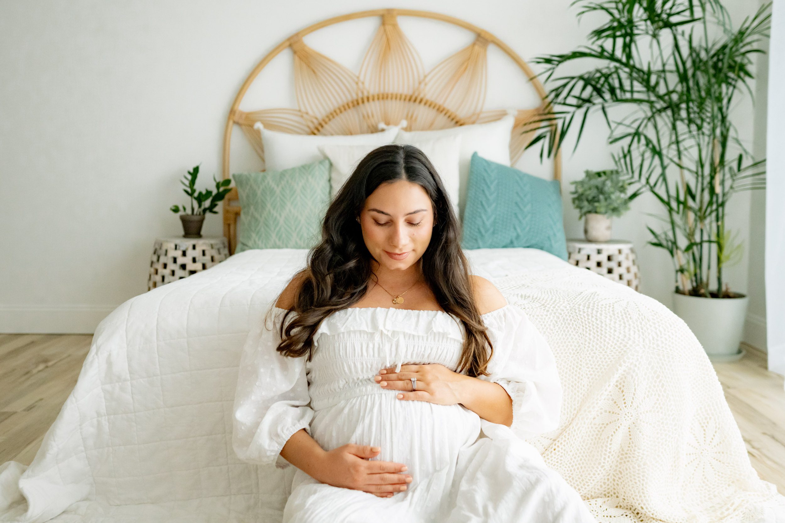 a pregnant woman dressed in a white off-shoulder gown is seen sitting in front of a bed, smiling affectionately at her belly during a lifestyle maternity photoshoot