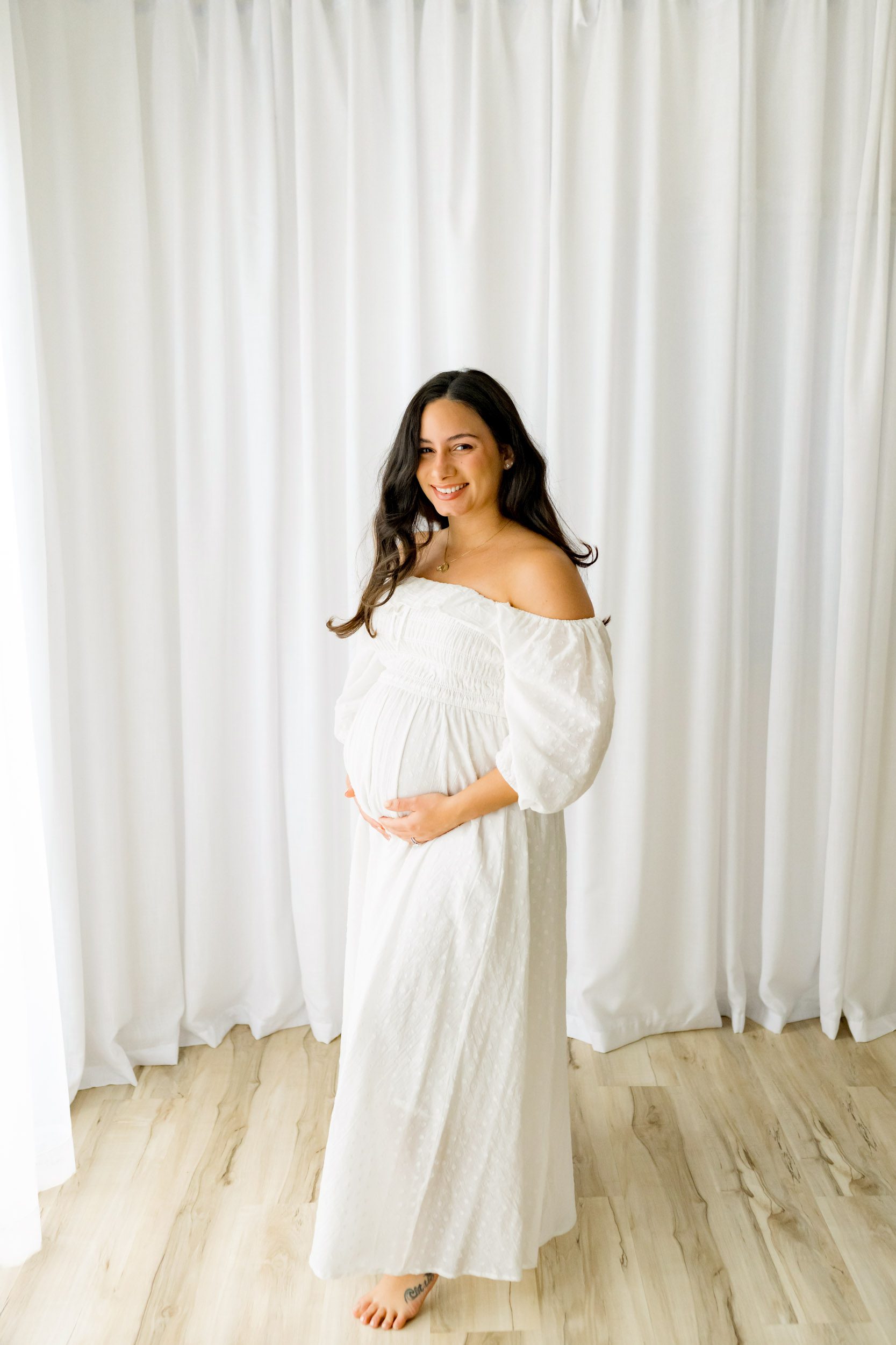 a pregnant woman dressed in a flowing white off-shoulder gown is positioned in front of a white curtain, beaming at the camera while she gently holds her belly during a lifestyle maternity photoshoot