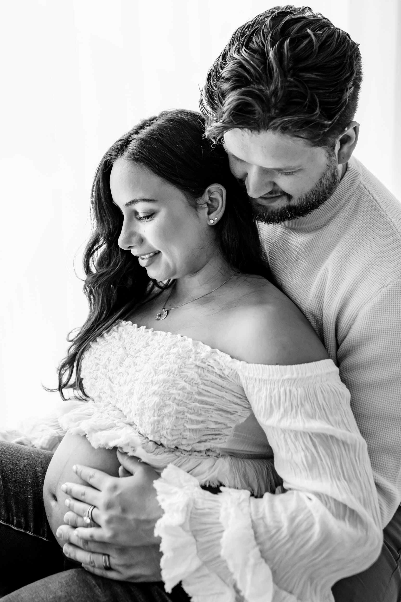 a black and white silhouette photograph of an expecting mother sporting a textured top and jeans seated on a bench with her husband behind her, both sharing smiles directed at her belly during a lifestyle maternity photoshoot