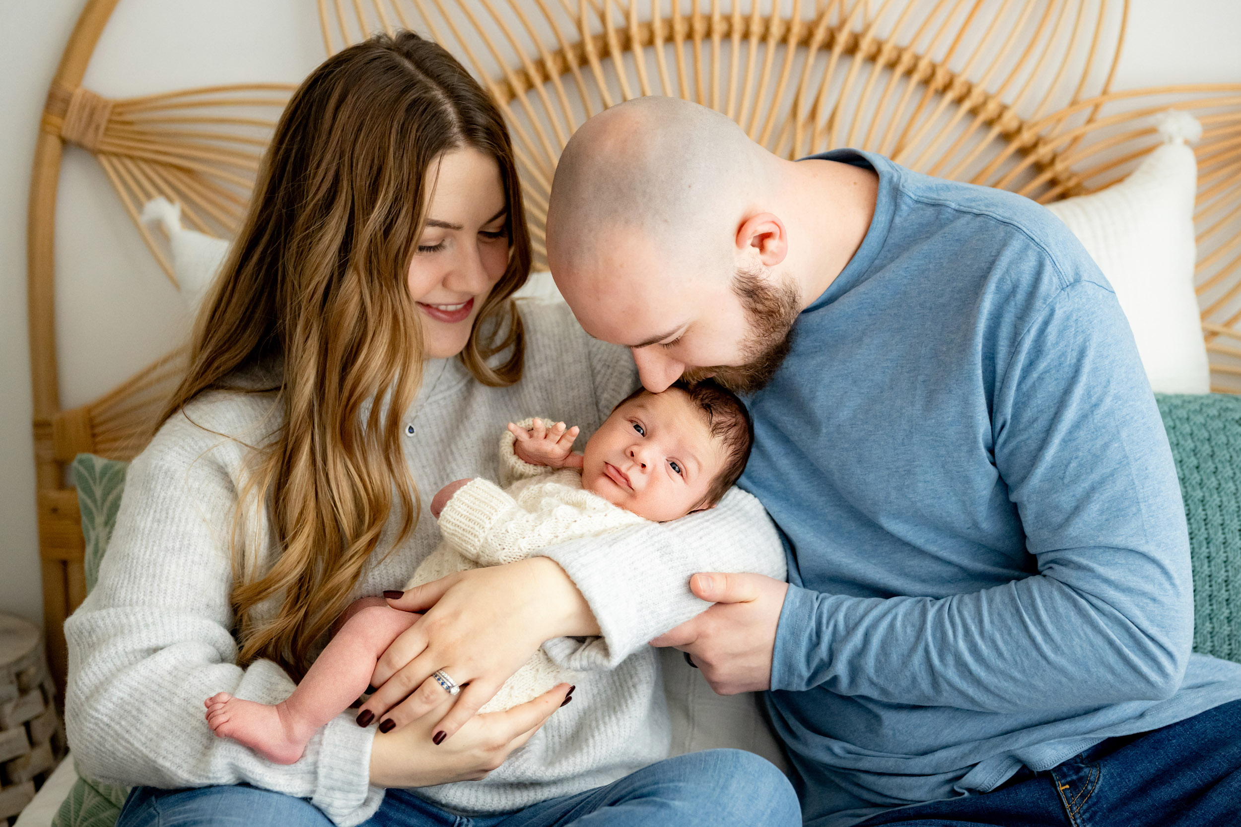 new parents seated on a bed with mom holding her baby boy in her arms while dad affectionately kisses him on the forehead as the baby looks straight at the camera during a newborn lifestyle photoshoot