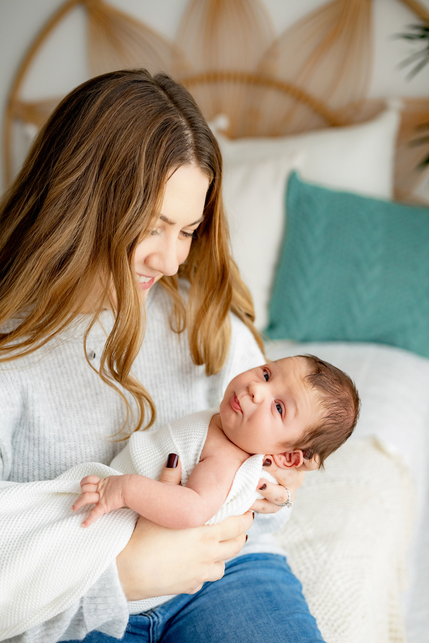 a new mother seated and smiling at her baby boy in her embrace as he purses his lips and gazes straight at the camera during a newborn lifestyle photoshoot