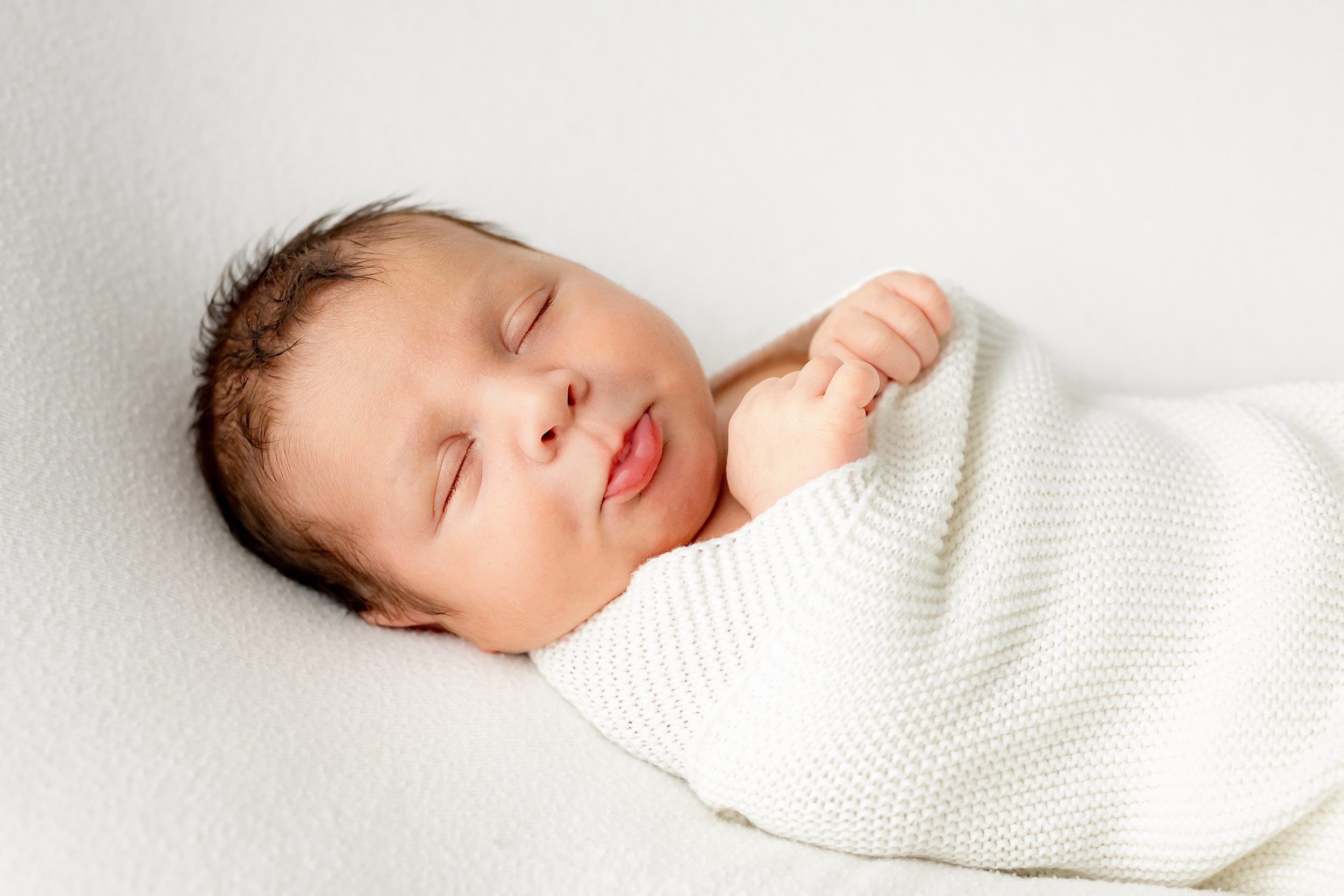 a baby boy swaddled in a white wrap resting on a white backdrop while puckering his lips in peaceful sleep during a newborn lifestyle photoshoot