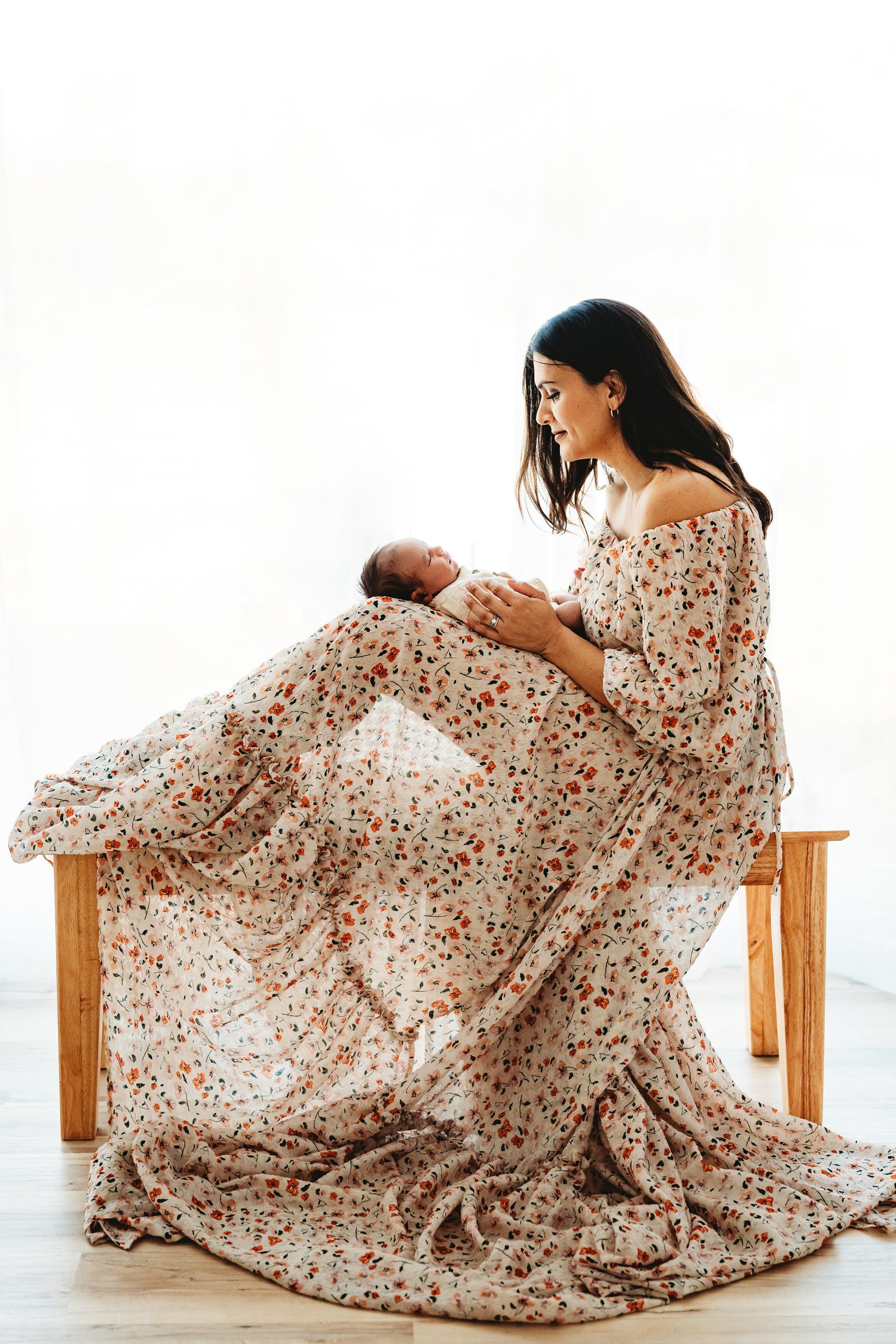 a backlit photo of a new mom wearing a long floral dress sitting on a bench holding her baby boy and smiling down at him during a newborn photoshoot