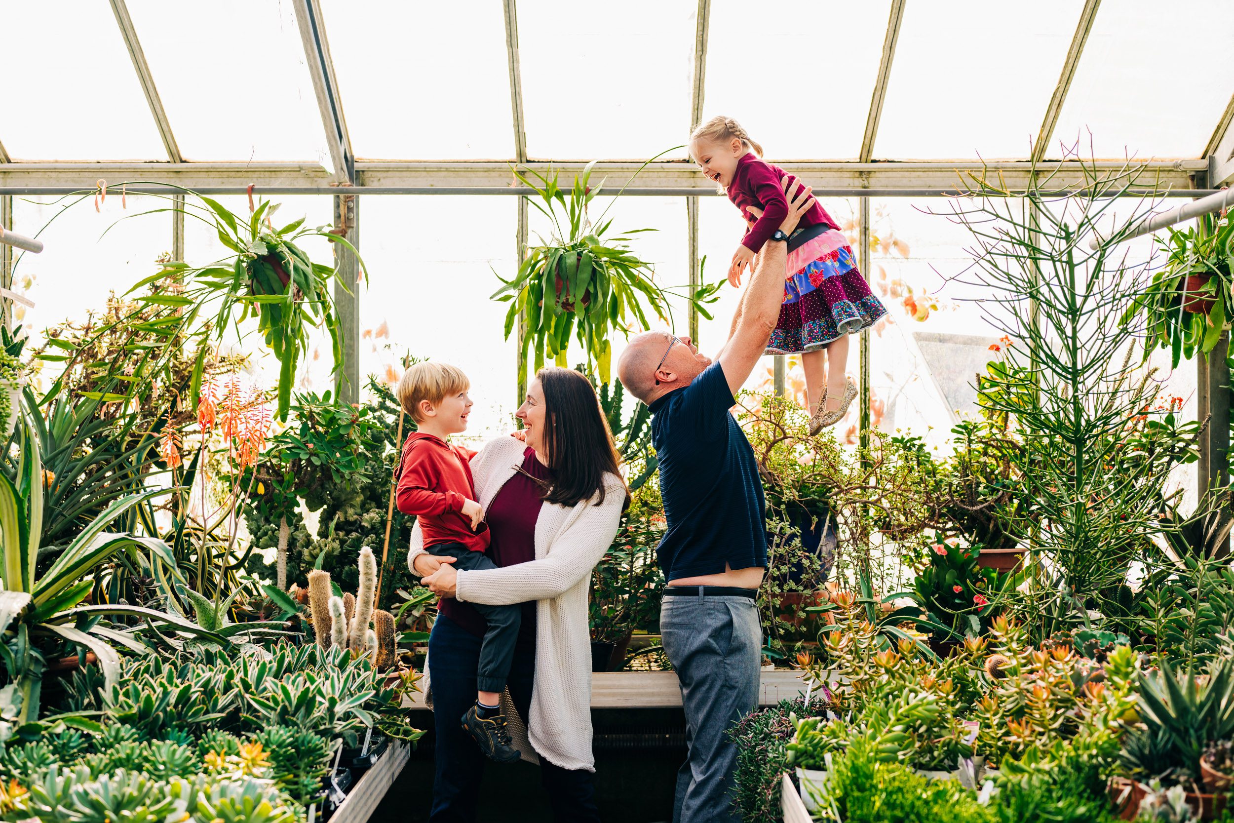 a family of four standing in a greenhouse as mom holds her son in her arms and dad lifts his daughter up in the air during a greenhouse family photoshoot