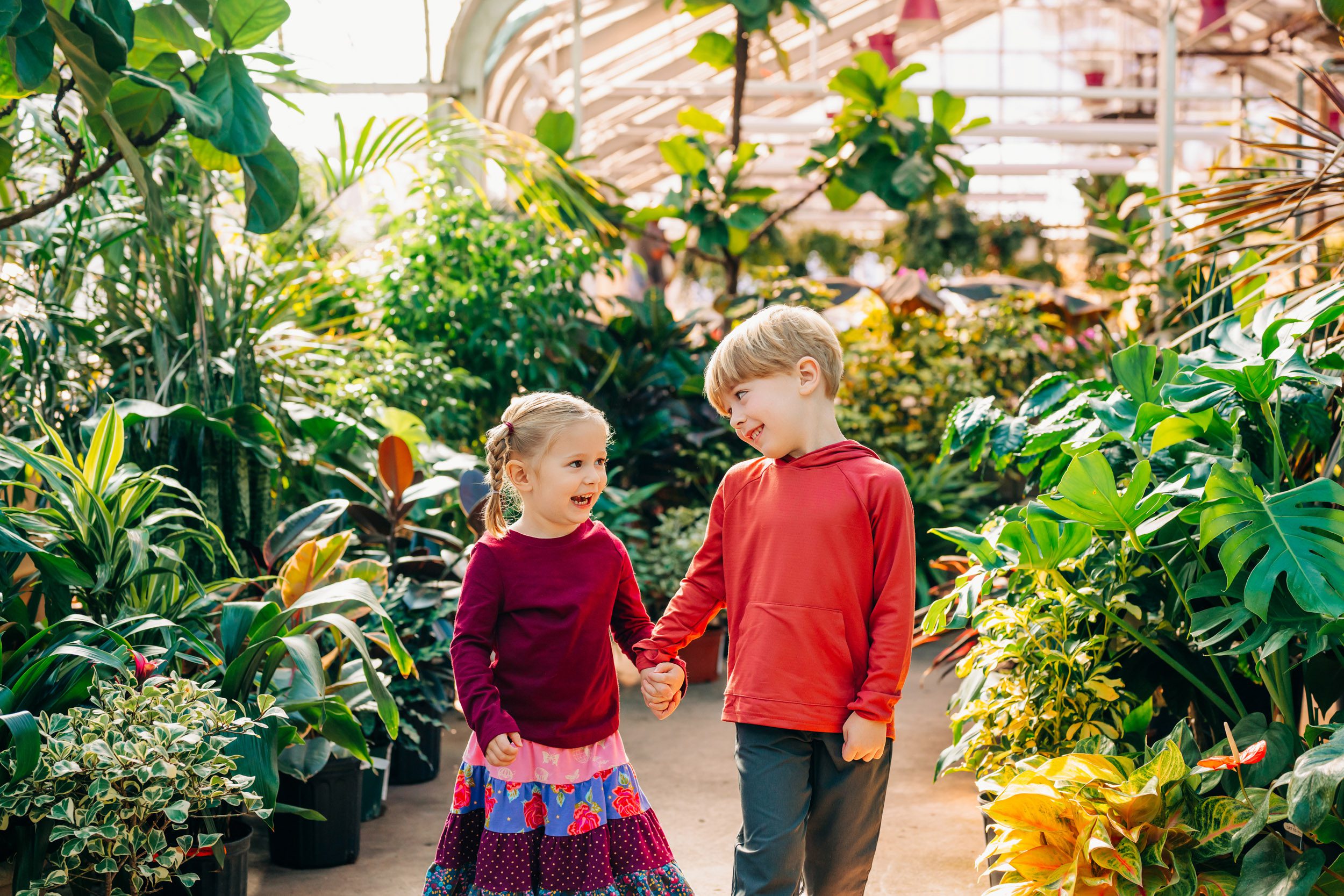 a young brother and sister holding hands and smiling at each other during a greenhouse family photoshoot