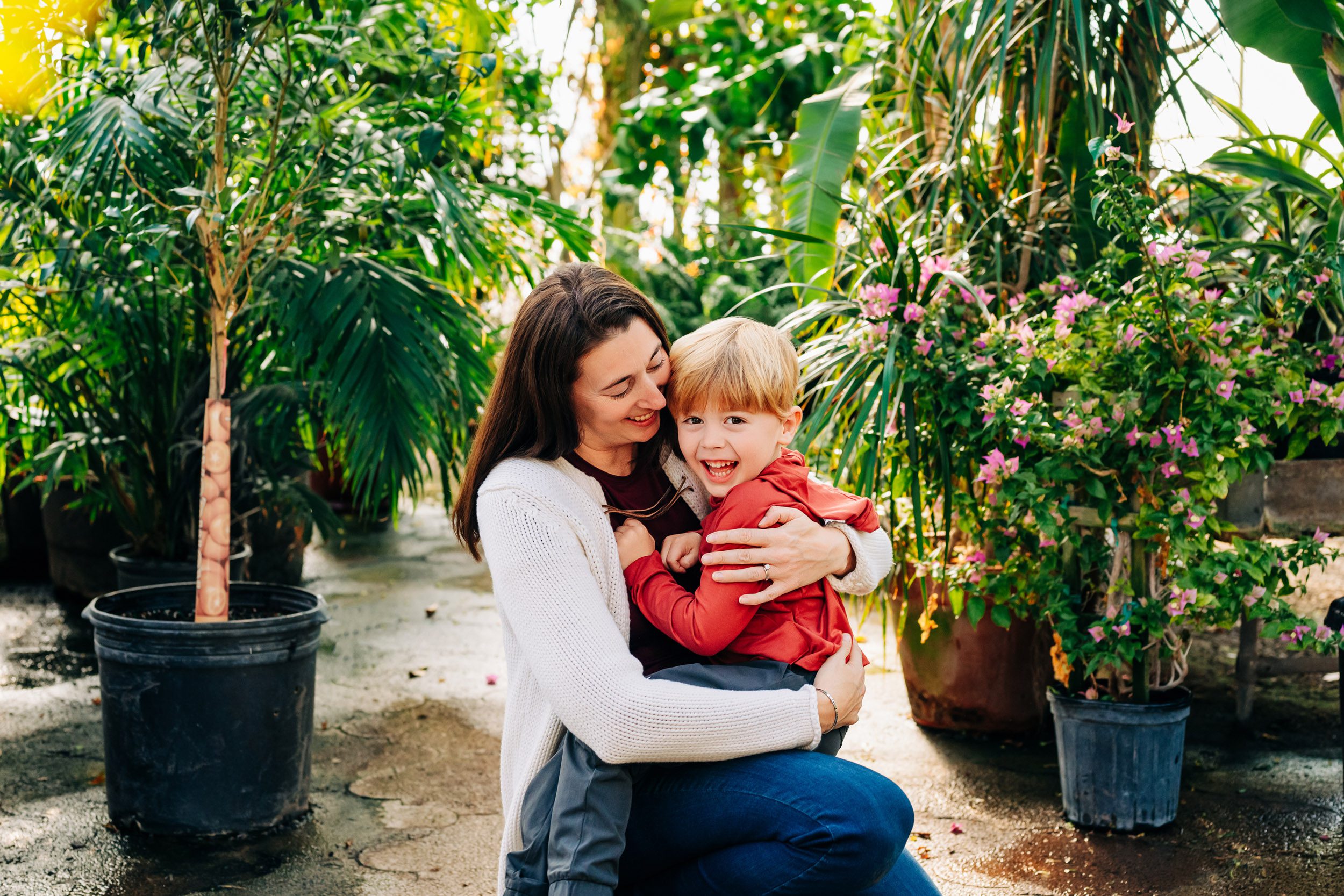 a mom kneeling down and hugging her son as he smiles at the camera during a greenhouse family photoshoot