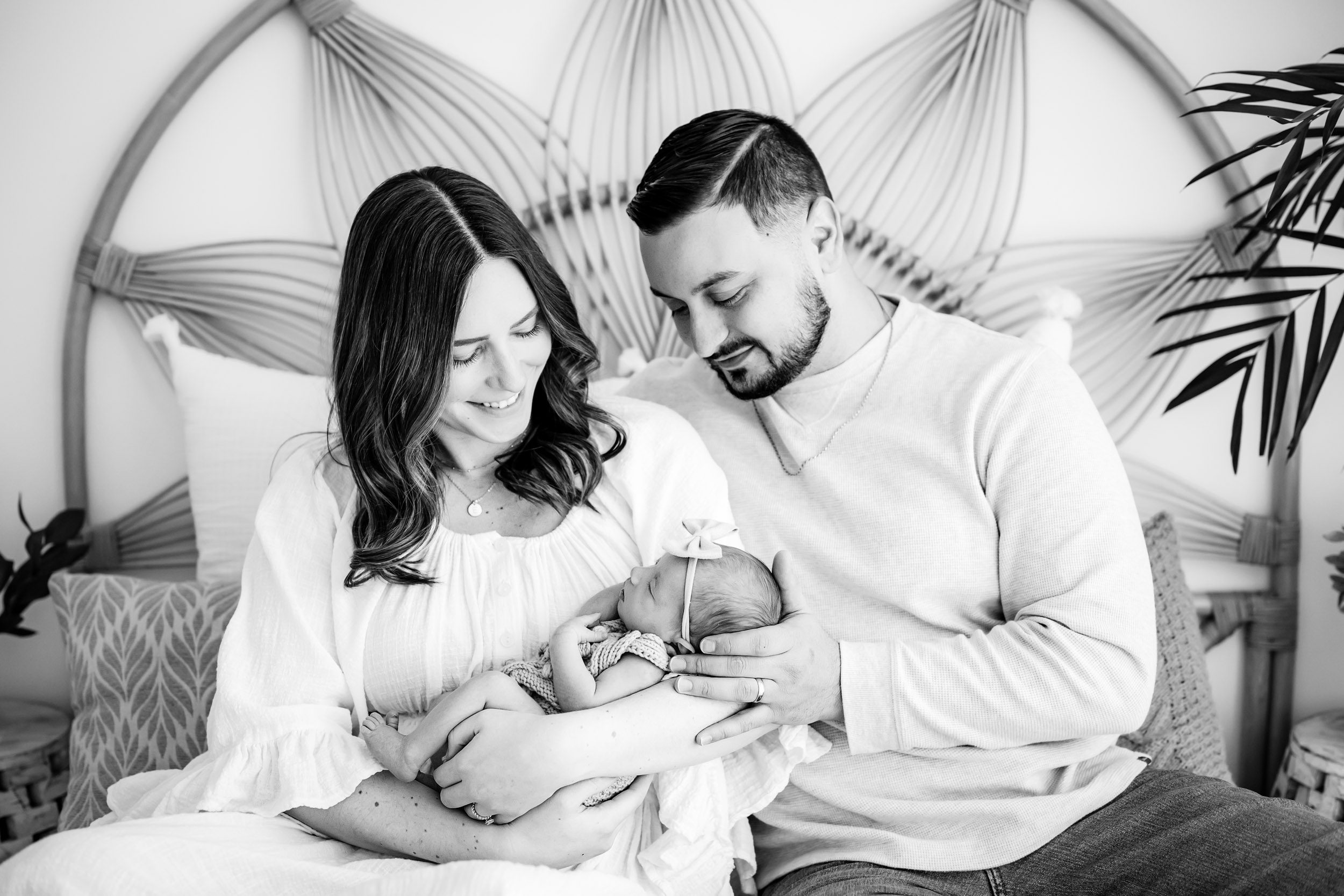 a black and white picture of parents sitting on a bed and cradling their baby girl in the arms as they smile down at her during a newborn baby photography session