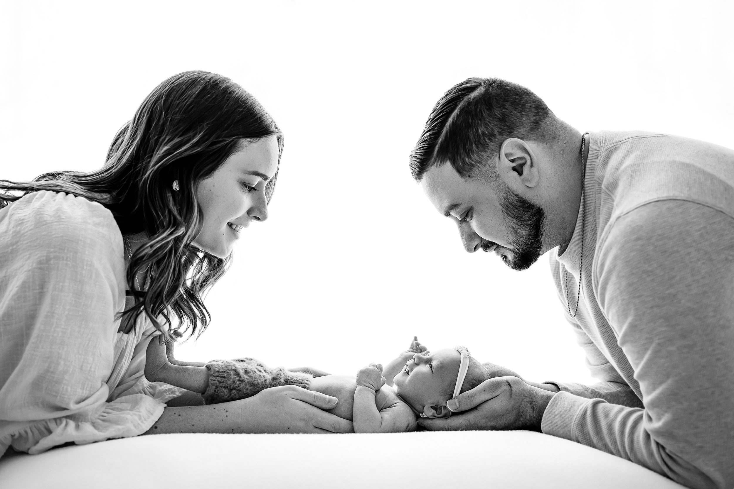 a black and white backlit photo of a baby girl laying on a white bean bag as her mom and dad smile down at her during a newborn baby photography session