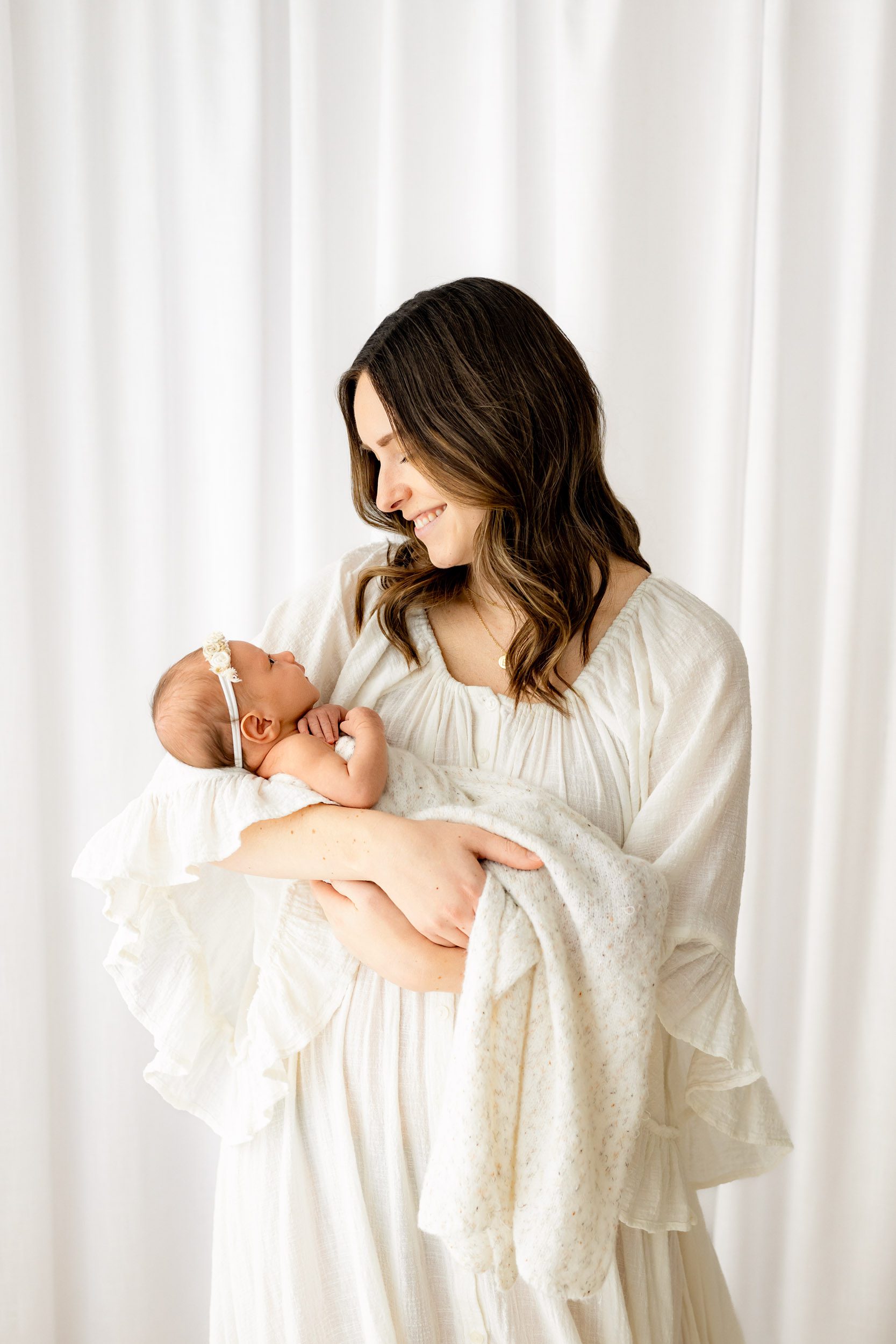 a new mother wearing a flowy white dress cradling her baby girl in her arms and smiling down at her during a newborn baby photography session