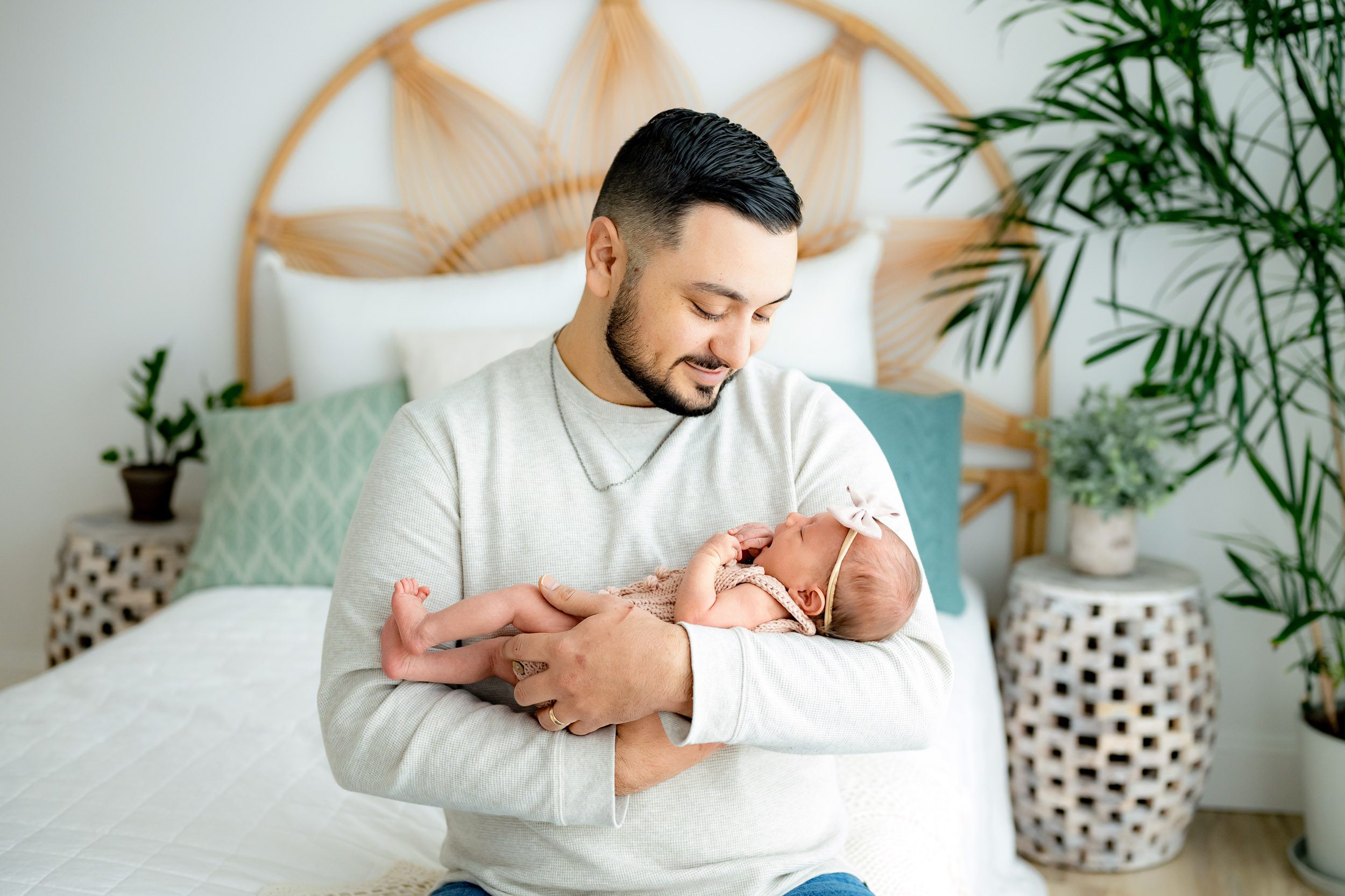 a new dad sitting on a bed and cradling his baby girl in his arms as he smiles down at her during a newborn baby photography session