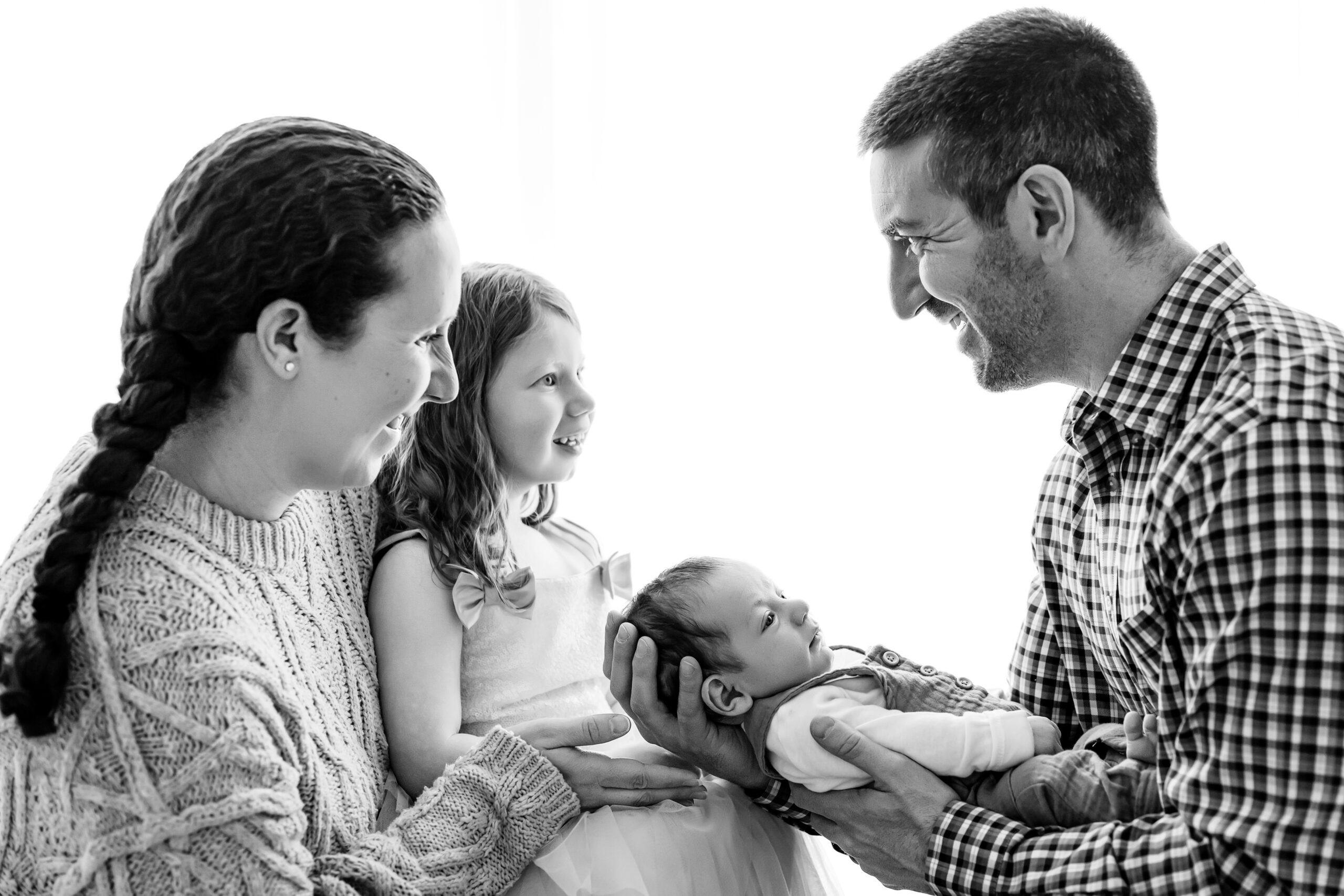 a black and white backlit picture of a dad holding his baby boy in his arms as mom holds her daughter on her lap and they smile at each other during a Pottstown newborn photography photoshoot