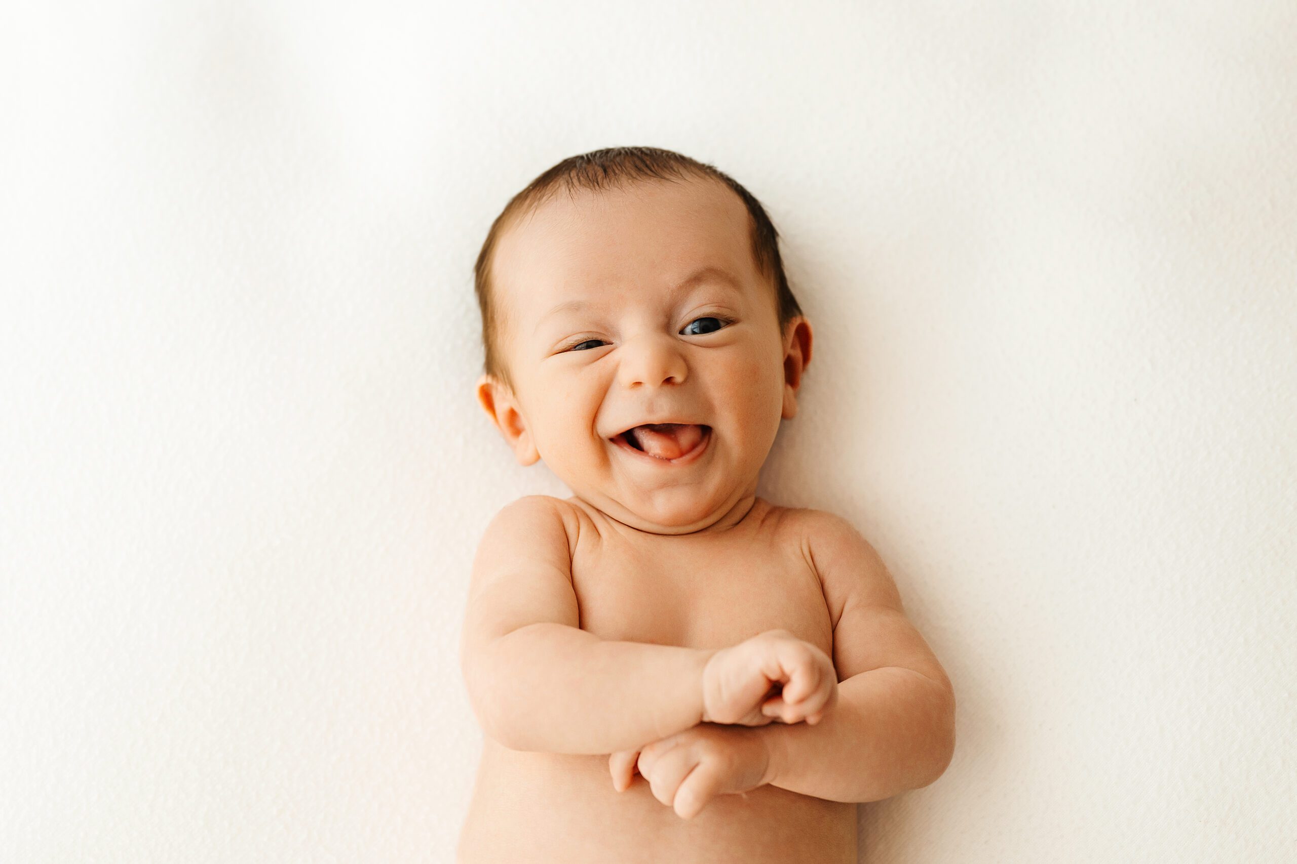 a baby boy laying on a white backdrop and sticking out his tongue with a silly smile on his face during a Pottstown newborn photography photoshoot