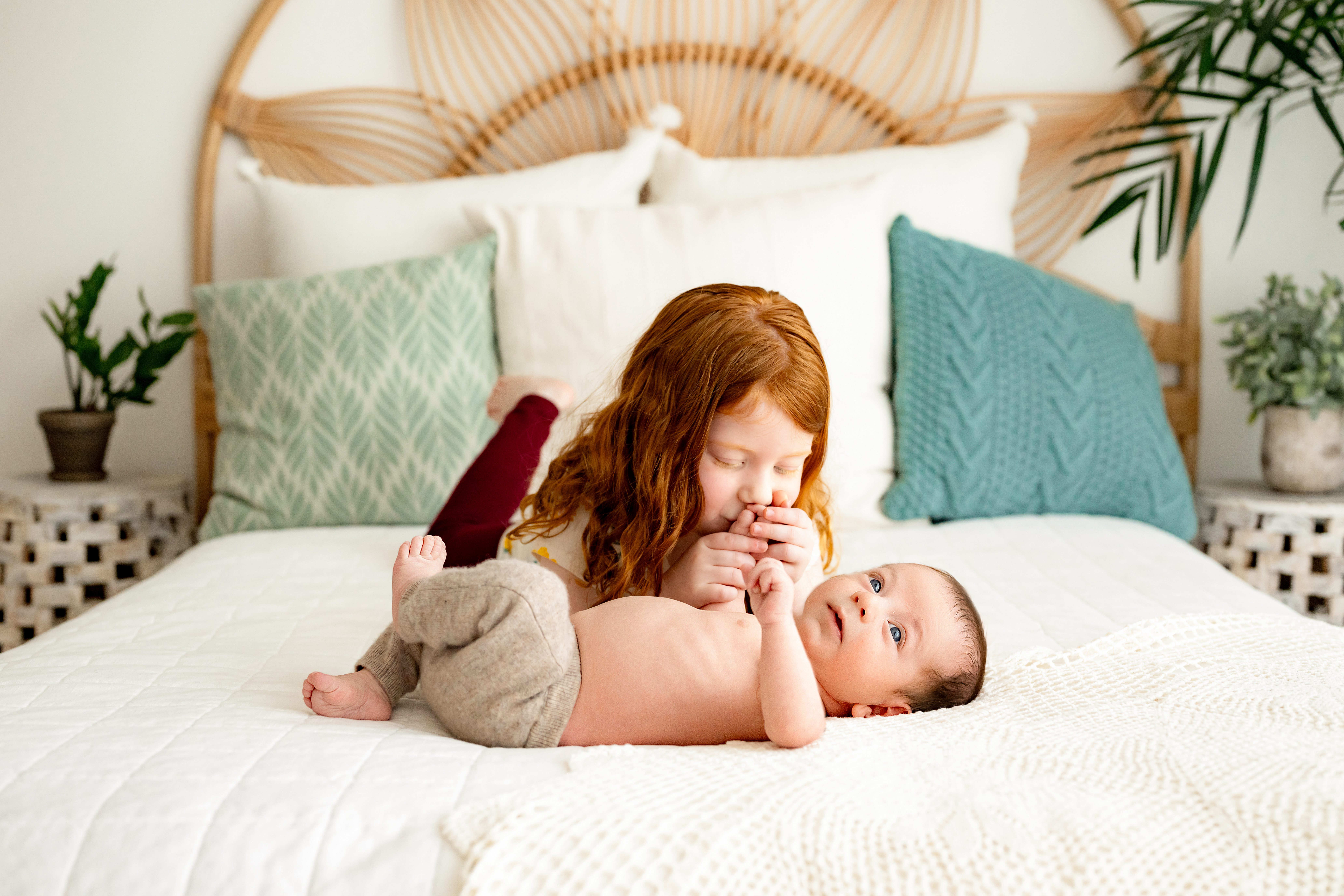 a baby boy laying on a bed with his big sister as she kisses his hand during a Pottstown newborn photography photoshoot