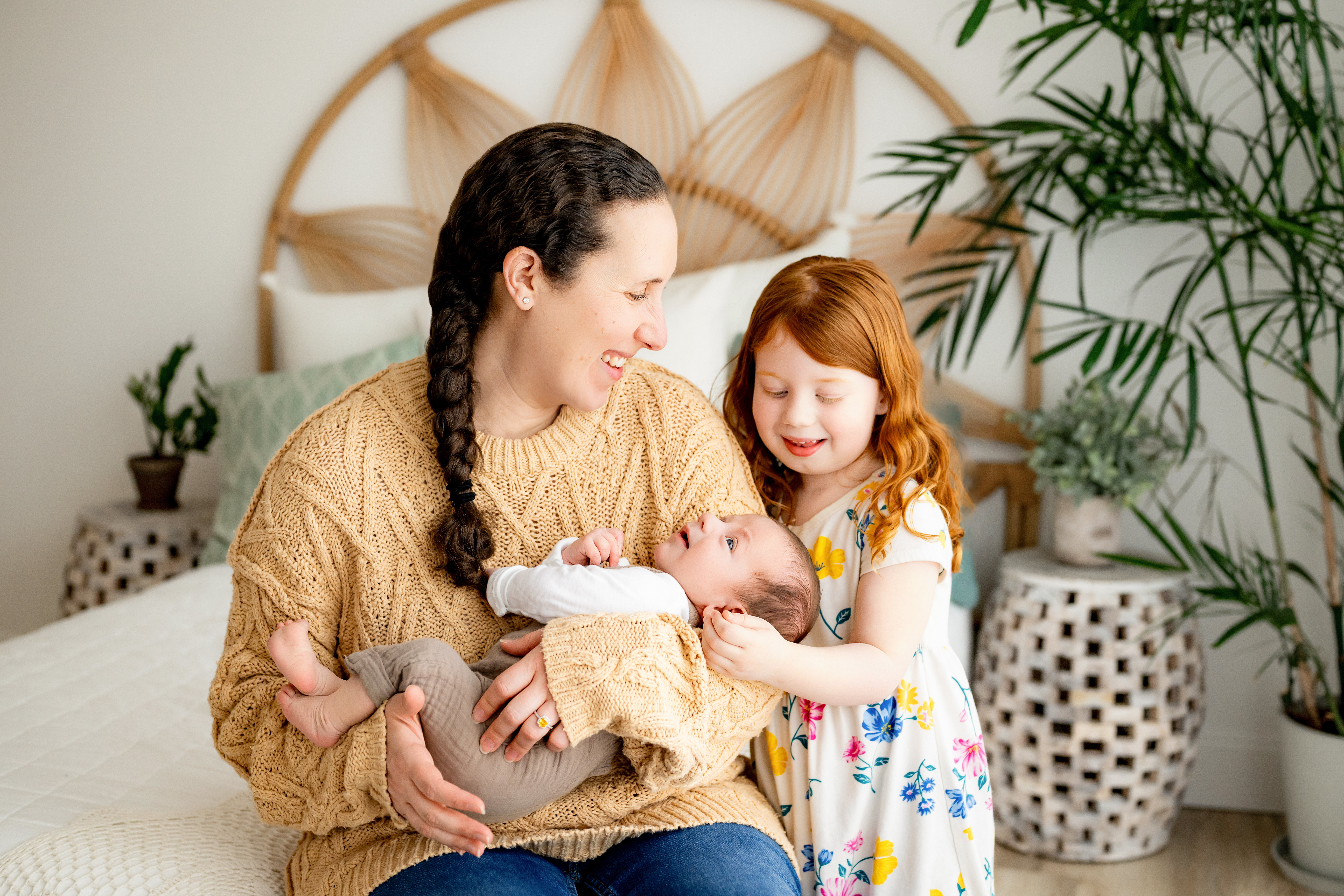 a mom sitting on a bed holding her baby boy in her arms as big sister touches her baby brother gently on the head and smiles down at him during a Pottstown newborn photography photoshoot
