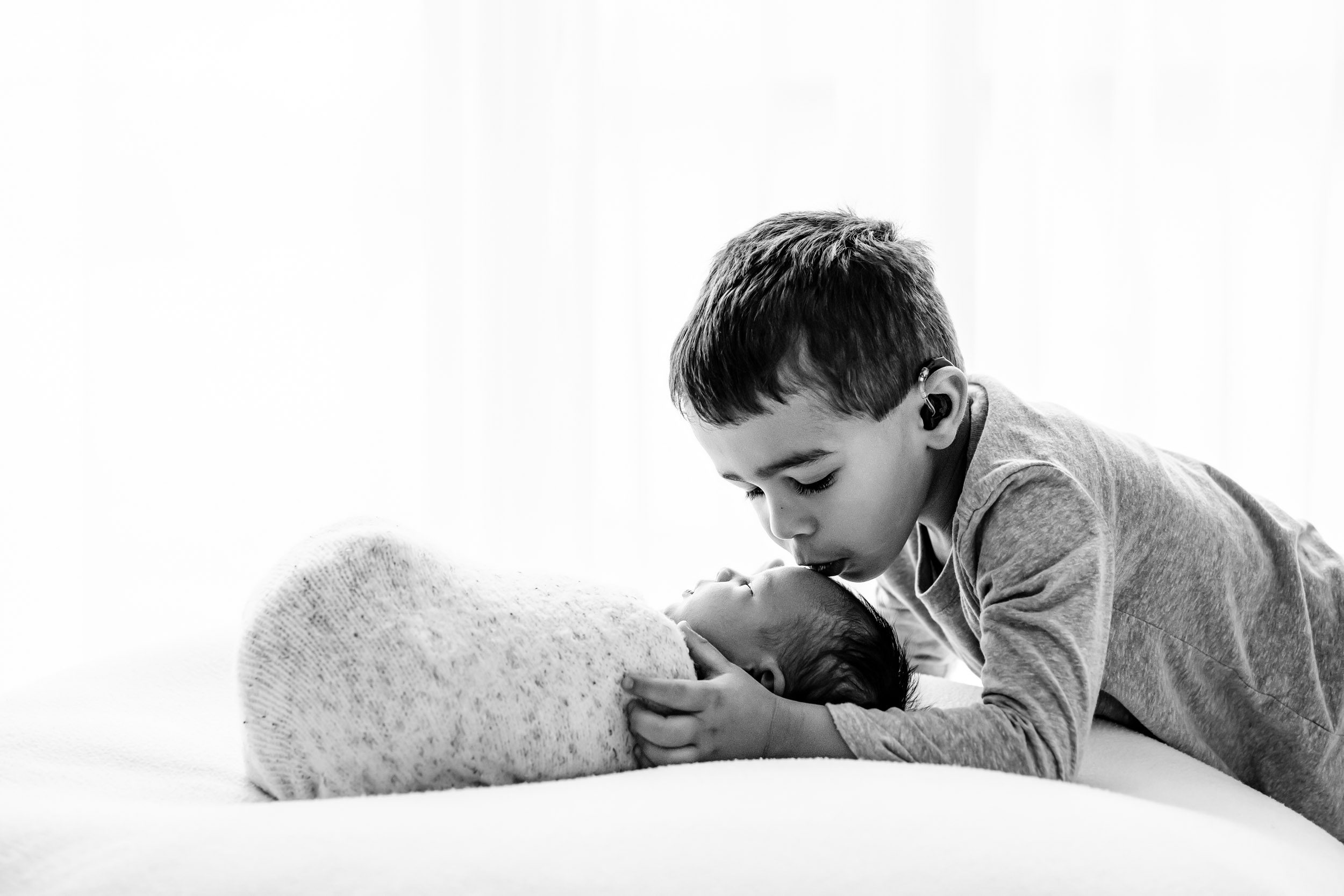 a black and white backlit picture of a baby boy laying on a bean bag with his sibling kisses him gently on the head during newborn photos