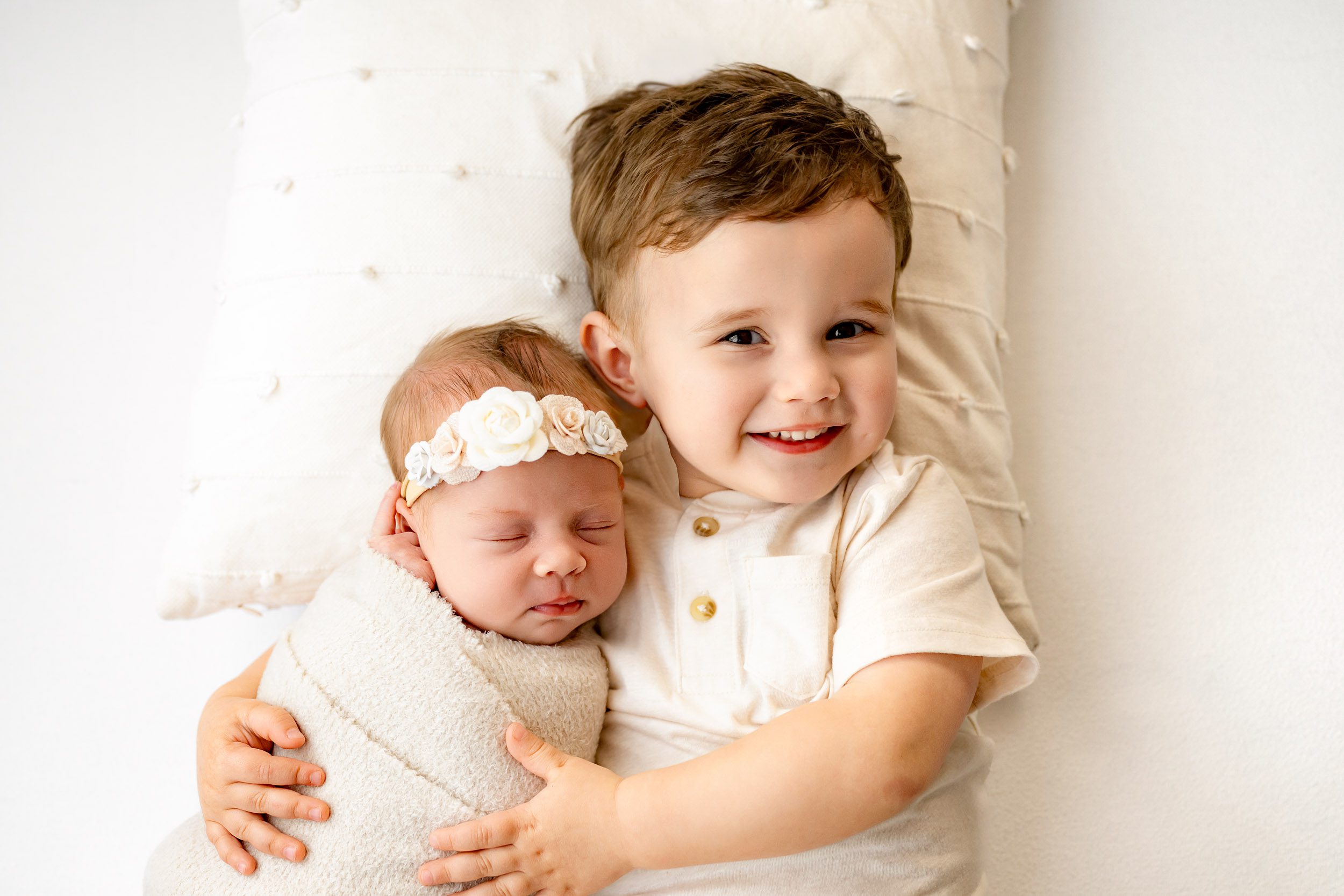 a picture taken from above of a big brother hugging his baby sister and smiling up at the camera during sibling newborn photos