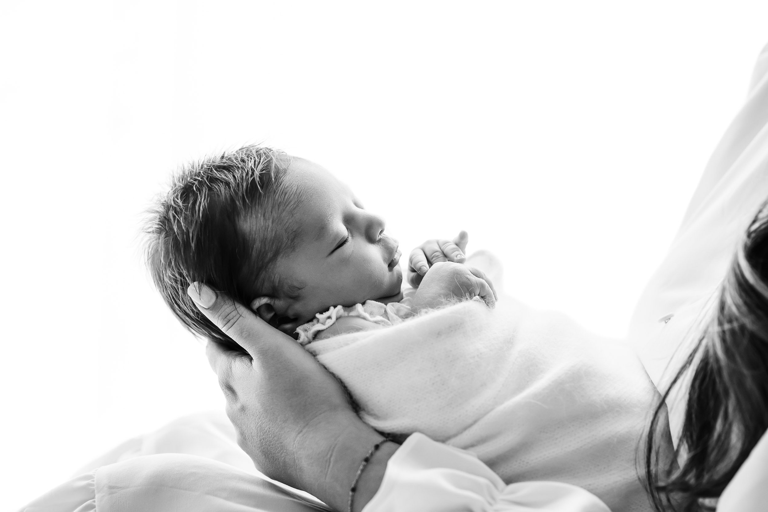 a black and white backlit photo of a baby girl wrapped in a fuzzy white swaddle sleeping peacefully in her mom's arms during a light and airy newborn photoshoot