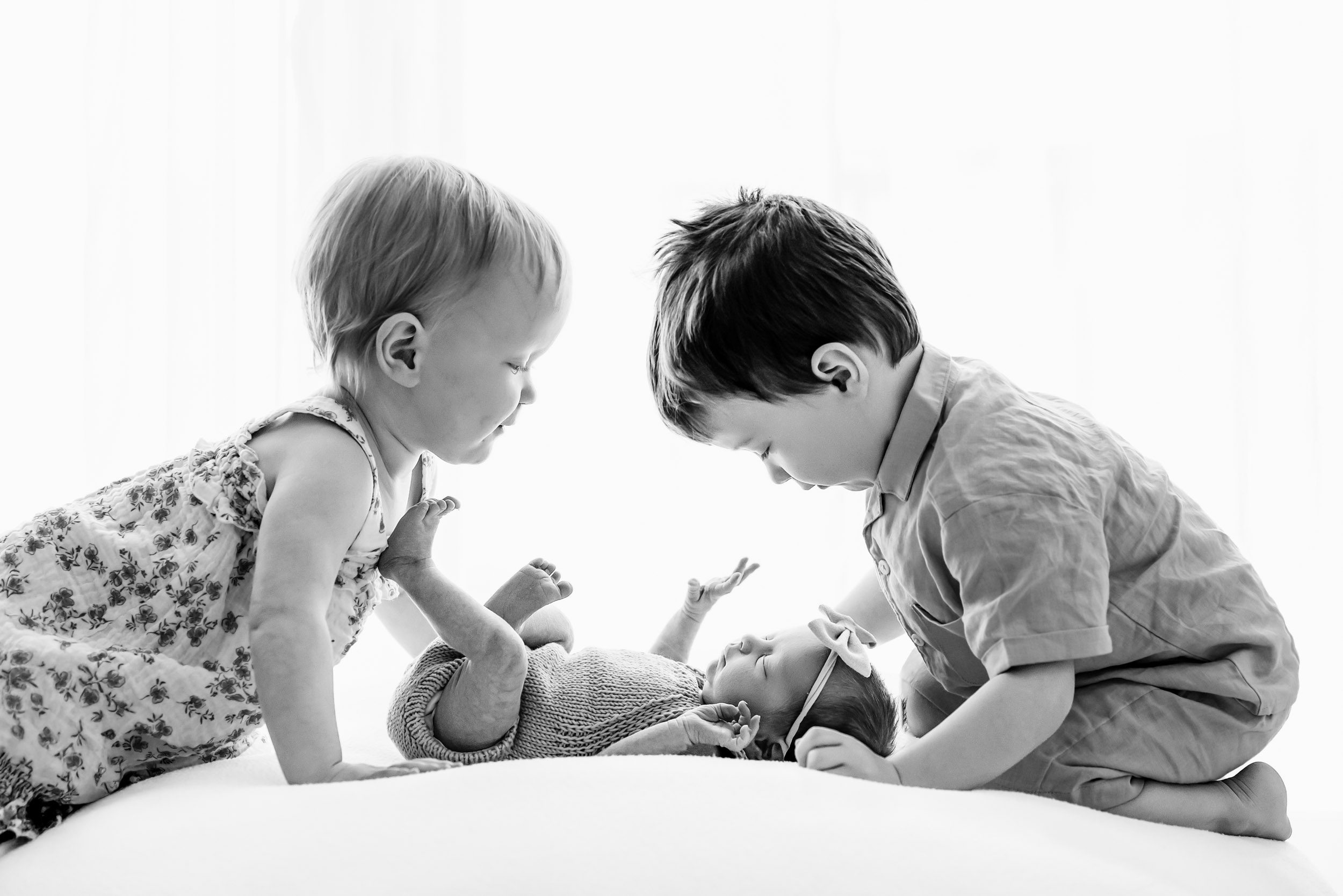 a black and white backlit baby picture of two young siblings smiling down at their baby sister as she lays on a bean bag