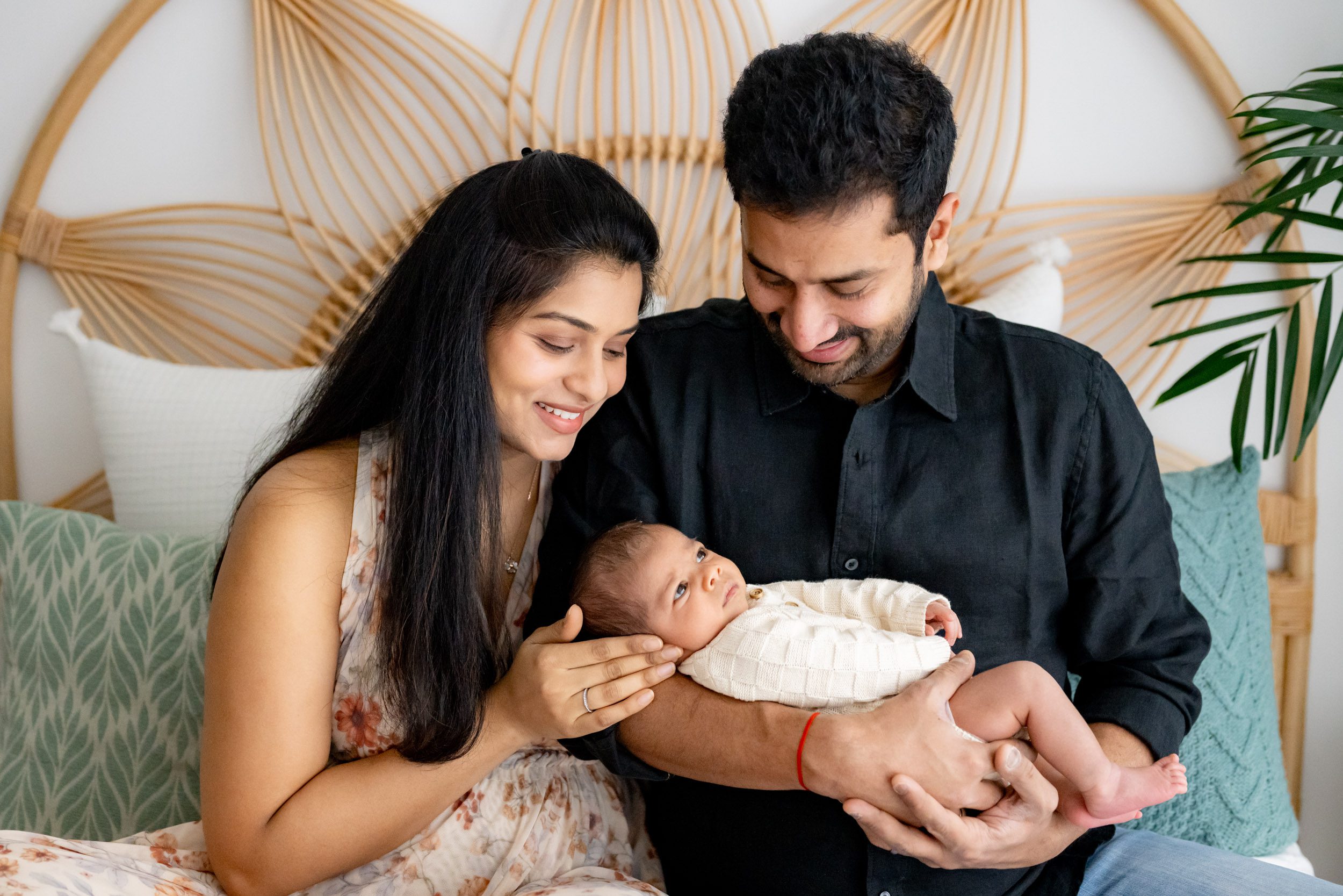 new parents sitting on a bed and smiling down at their baby boy cradled in dad's arms during a newborn photoshoot