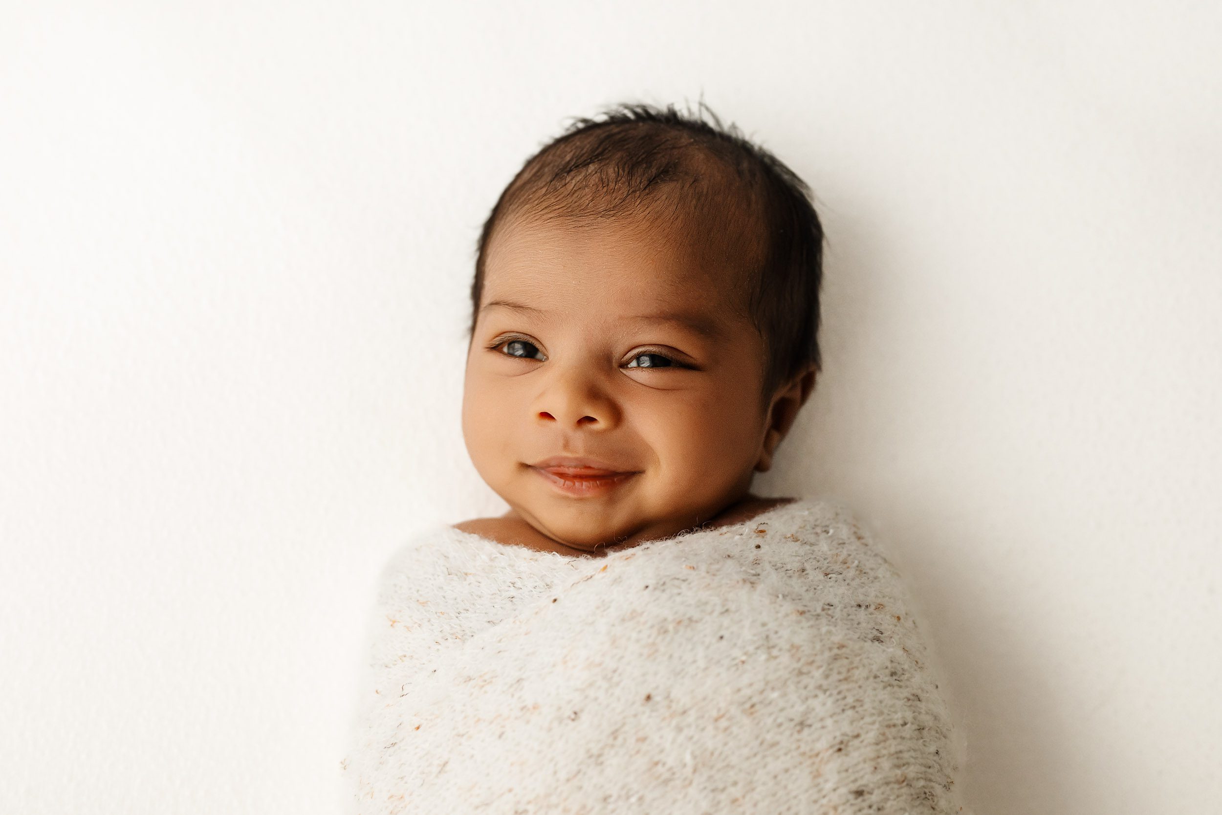 a baby boy wrapped in a fuzzy white swaddle blanket with speckles and gazing up with a hint of a smile on his face during a baby photoshoot