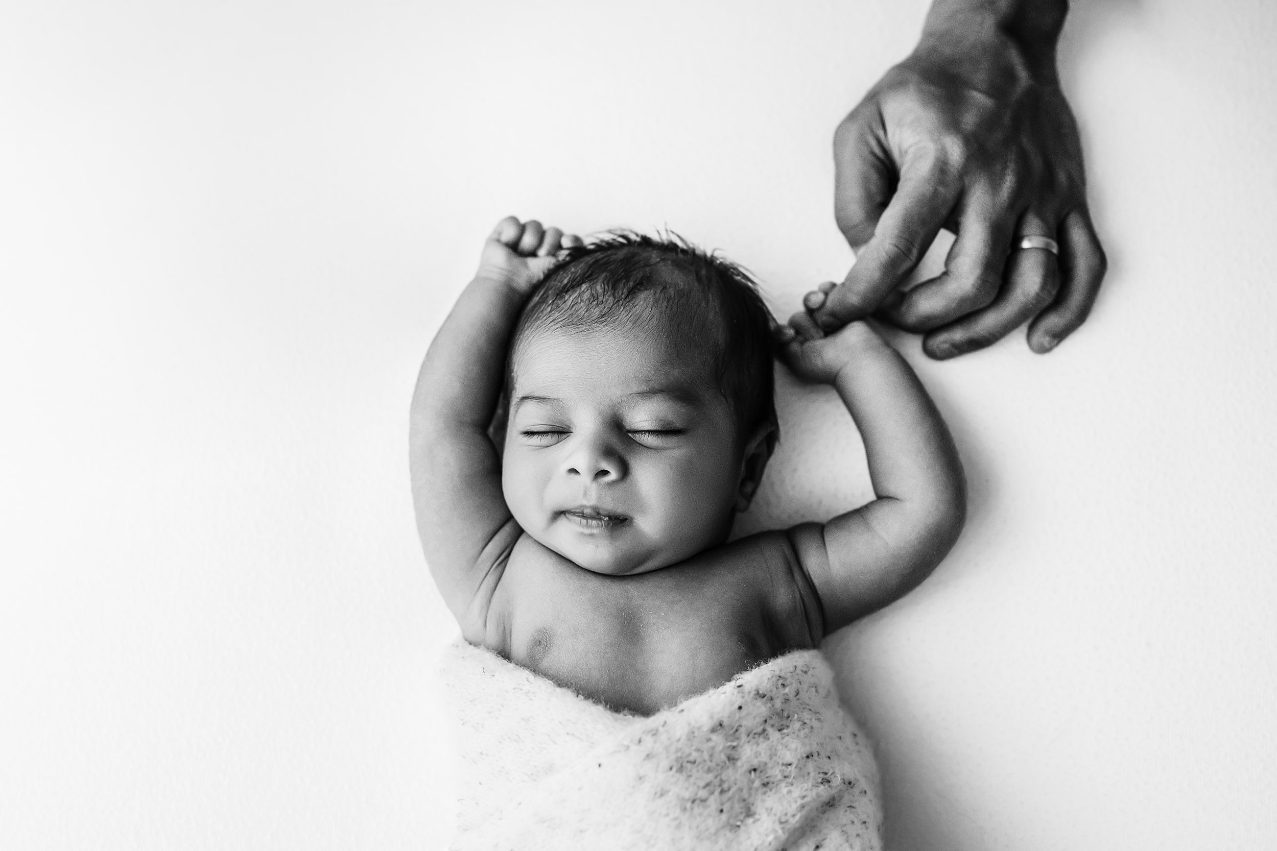 a black and white baby photo of a baby boy wrapped in a white swaddle blanket and sleeping peacefully as he holds his dad's finger with one of his hands