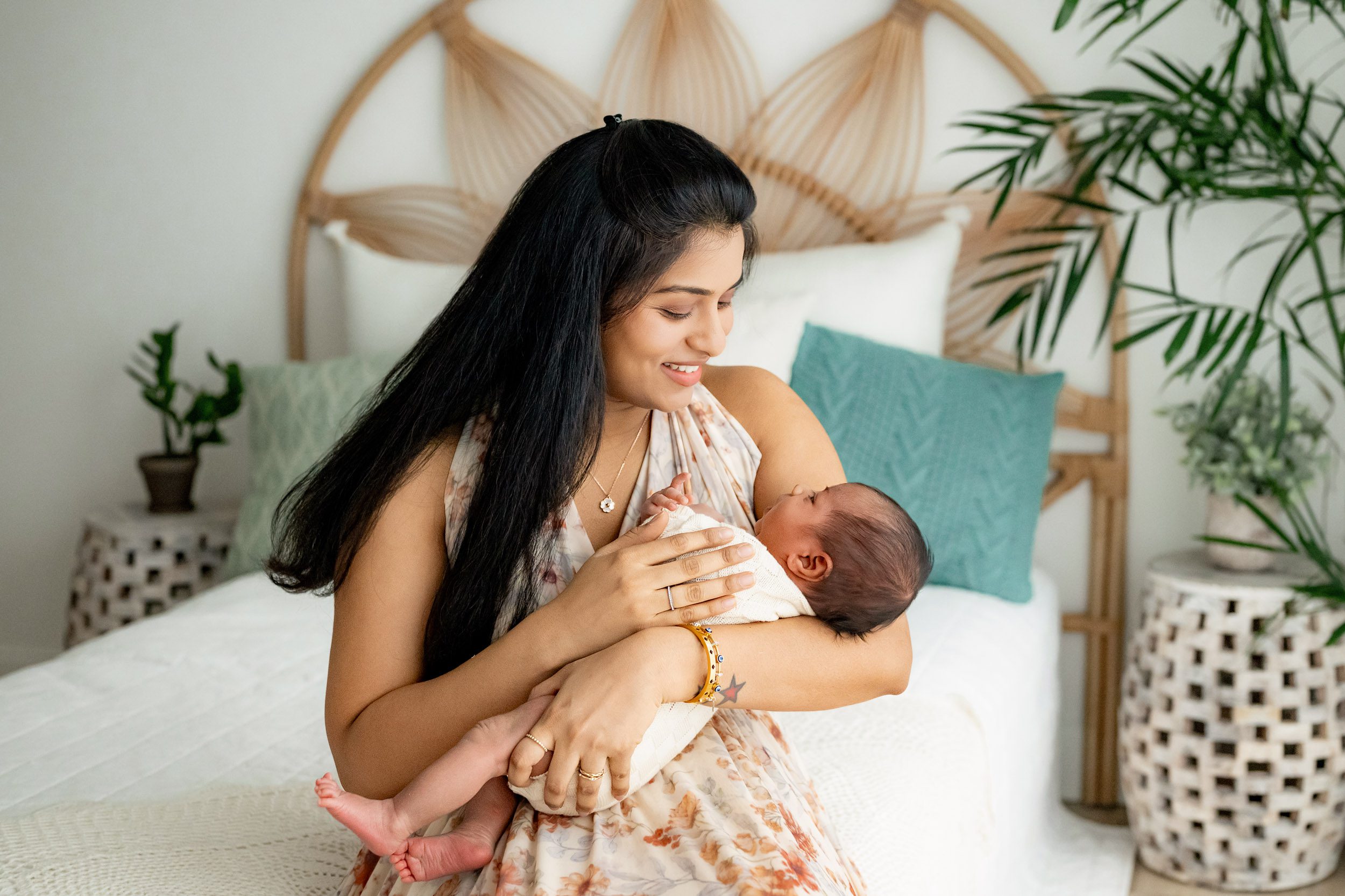 a new mother sitting on a bed smiling down at her baby boy cradled in her arms during a baby photoshoot