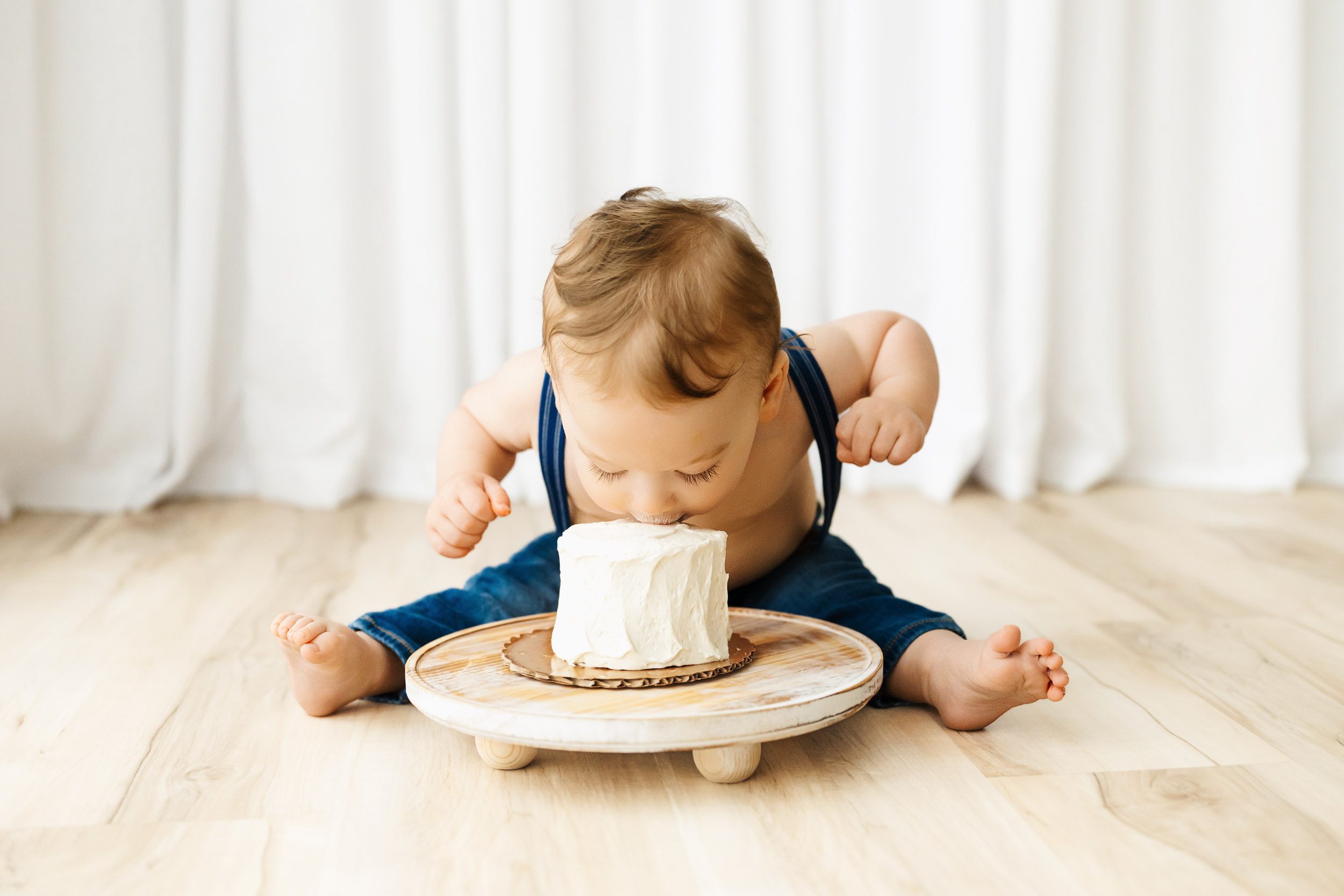 a young boy wearing blue jeans with suspenders sitting on the floor and leaning down to take a big bite out of his birthday cake during a 1st birthday photography session