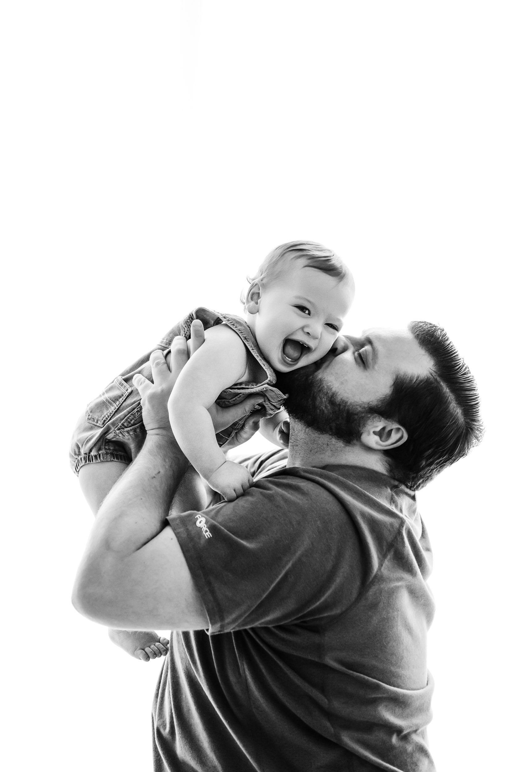 a black and white backlit photo of a dad kissing his baby boy on the cheek as he looks directly at the camera and laughs during a 1st birthday photography session