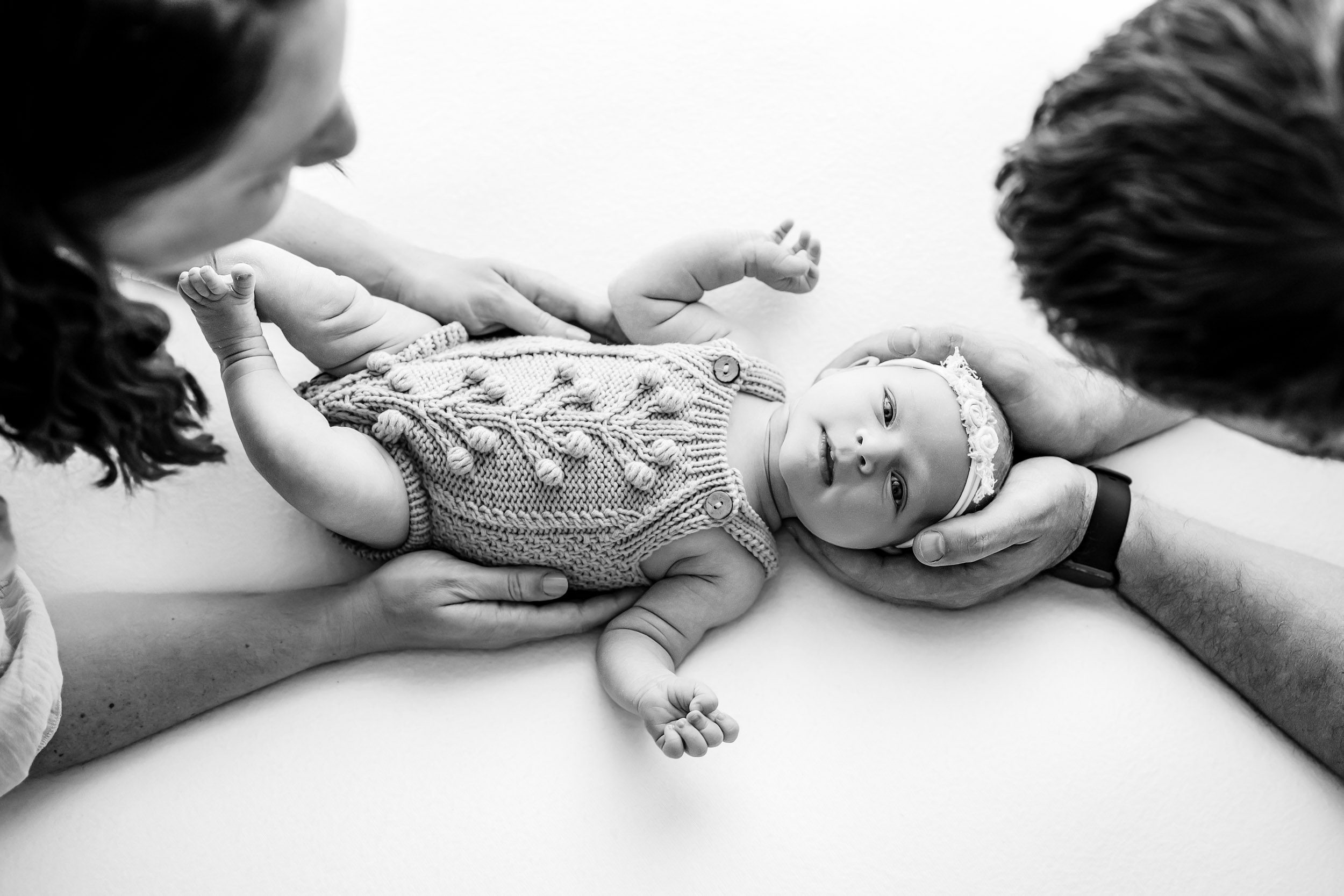 a black and white picture taken from above of a baby girl looking directly at the camera as her mom and dad put their hands around her during a newborn family photoshoot