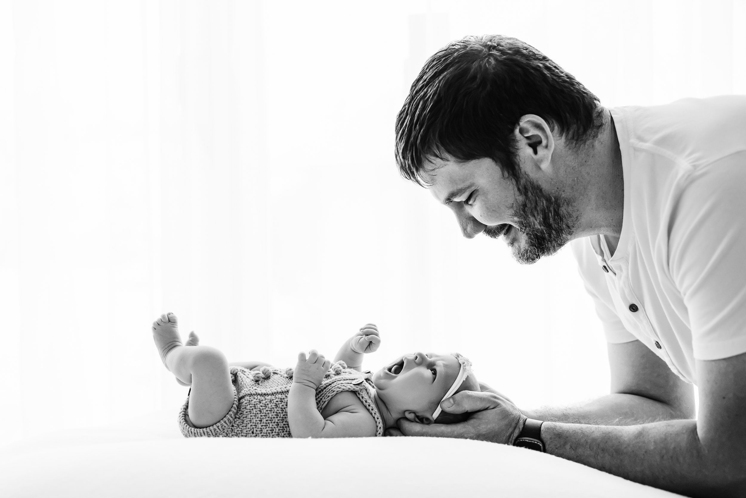 a black and white backlit photo of a baby girl laying on a white bean bag as her dad smiles down at her during a newborn family photoshoot