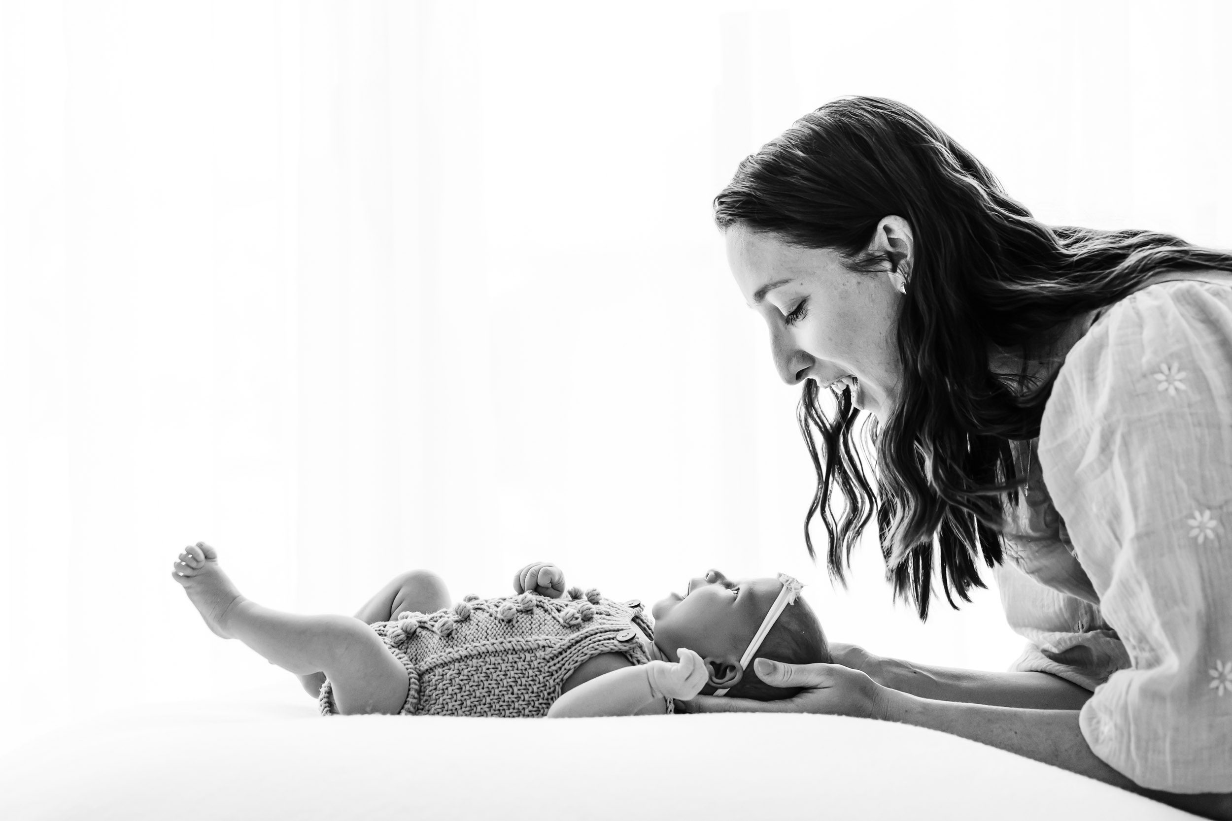 a black and white backlit photo of a baby girl laying on a white bean bag as her mom smiles down at her during a newborn family photoshoot